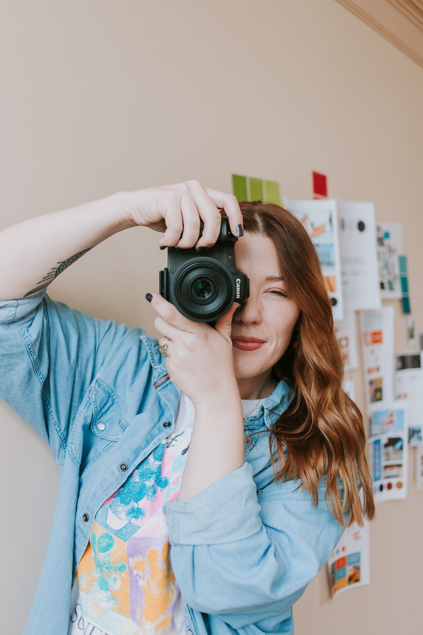 Young woman with auburn hair holding a Canon camera up to her eye, wearing a denim shirt, standing near a mood board.