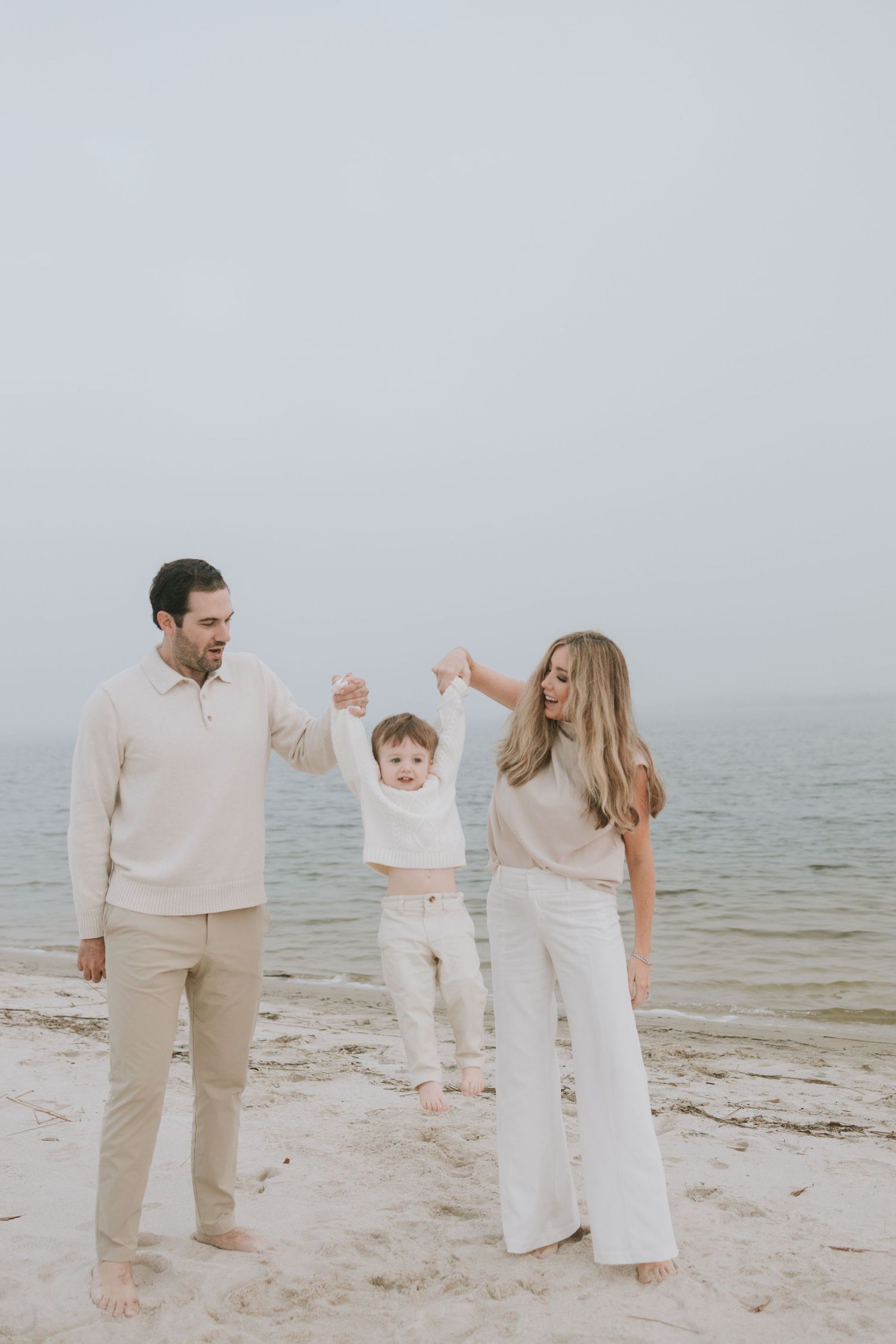 A young family in neutral tones holds their toddler's hands on a misty beach, lifting the child joyfully.