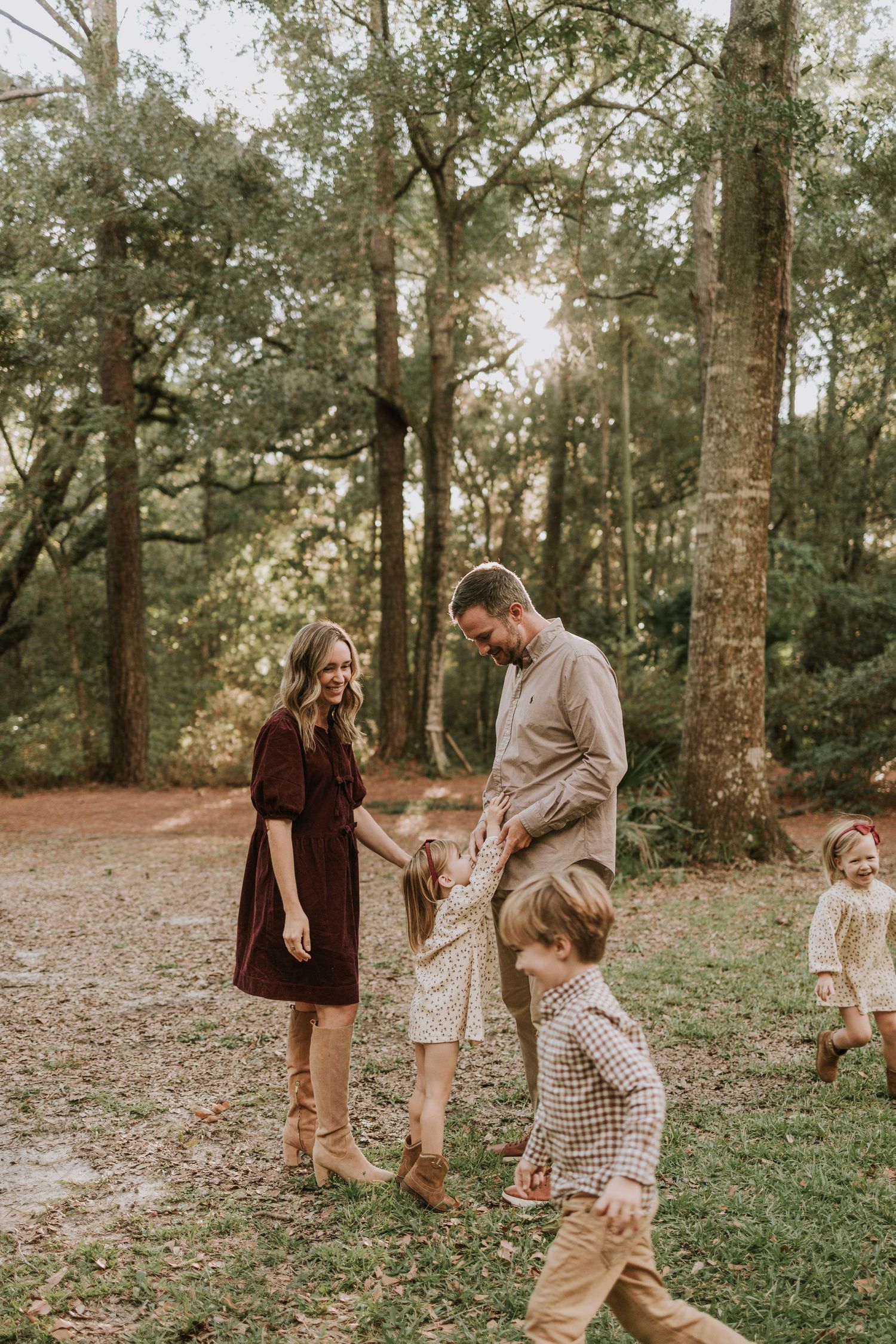 Family of four enjoying a sunny outdoor photo session among tall trees, parents smiling at young children playing.