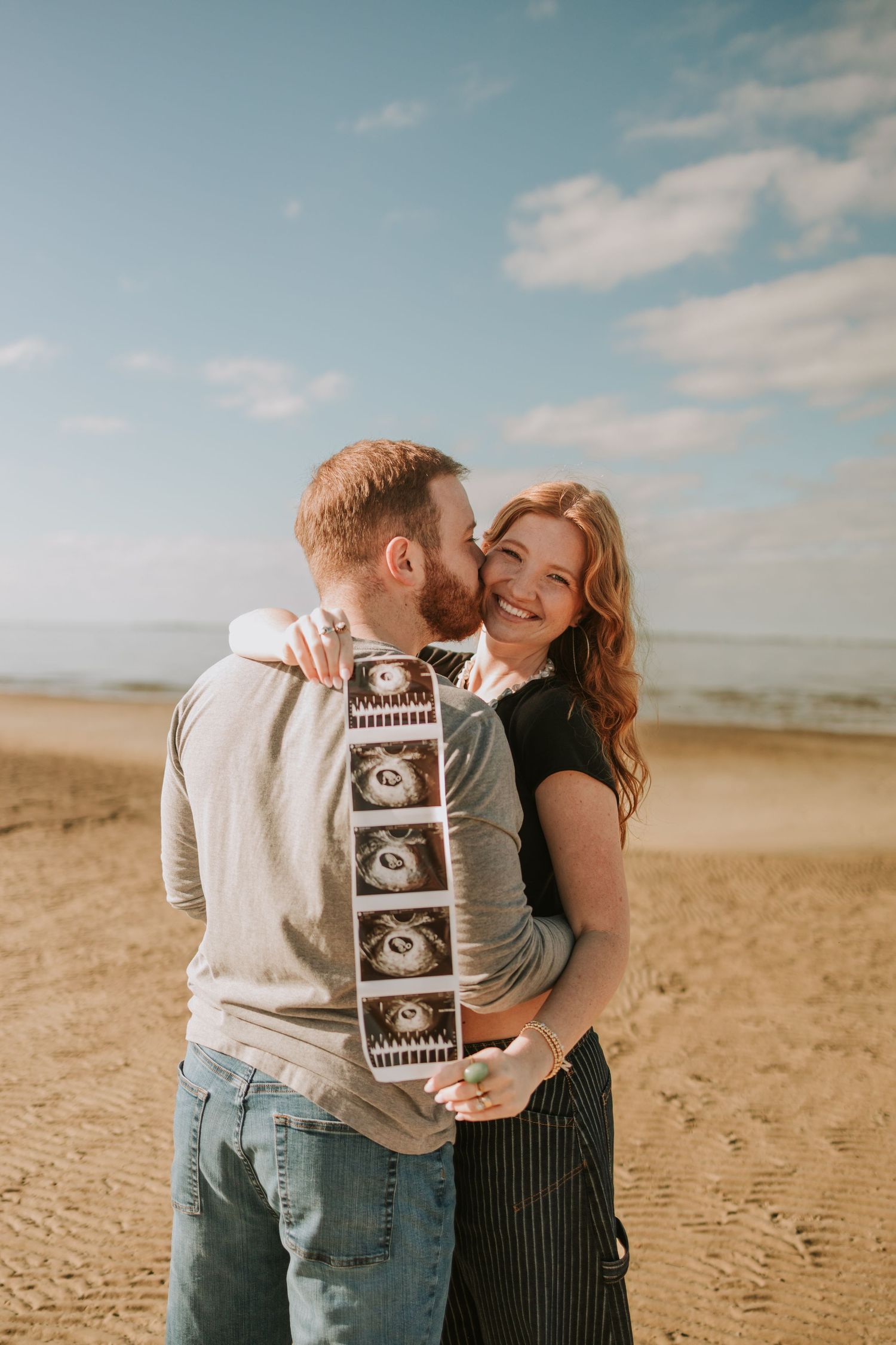 Couple embracing on sandy beach holding ultrasound photos, announcing pregnancy with joy and smiles.