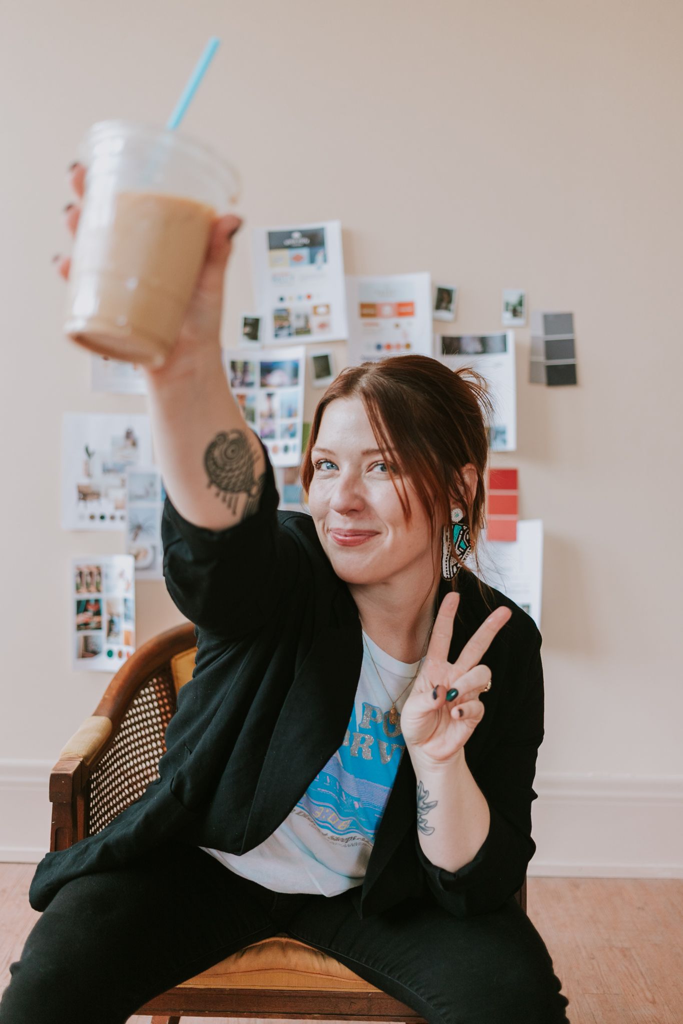 Woman with tattoos flashing peace sign and holding iced coffee, wearing black blazer and teal earrings, seated indoors.