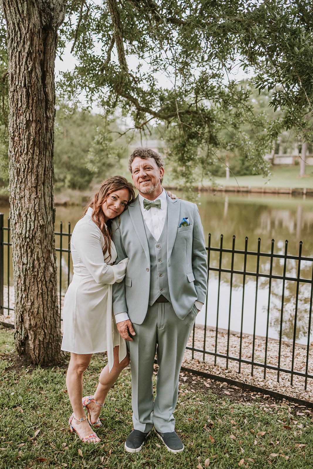 A couple poses together outdoors by a lake under a large tree with hanging branches and wrought iron railings.