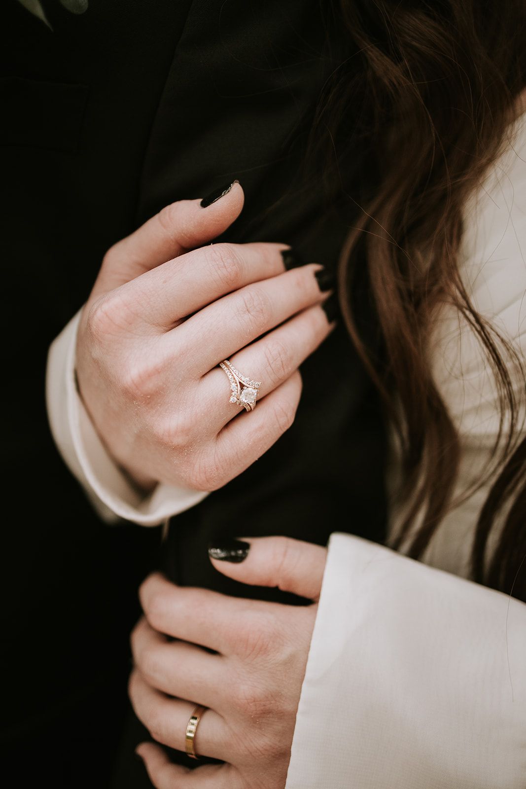 Hands with dark nail polish wearing delicate gold rings against a cream colored sleeve and dark background.