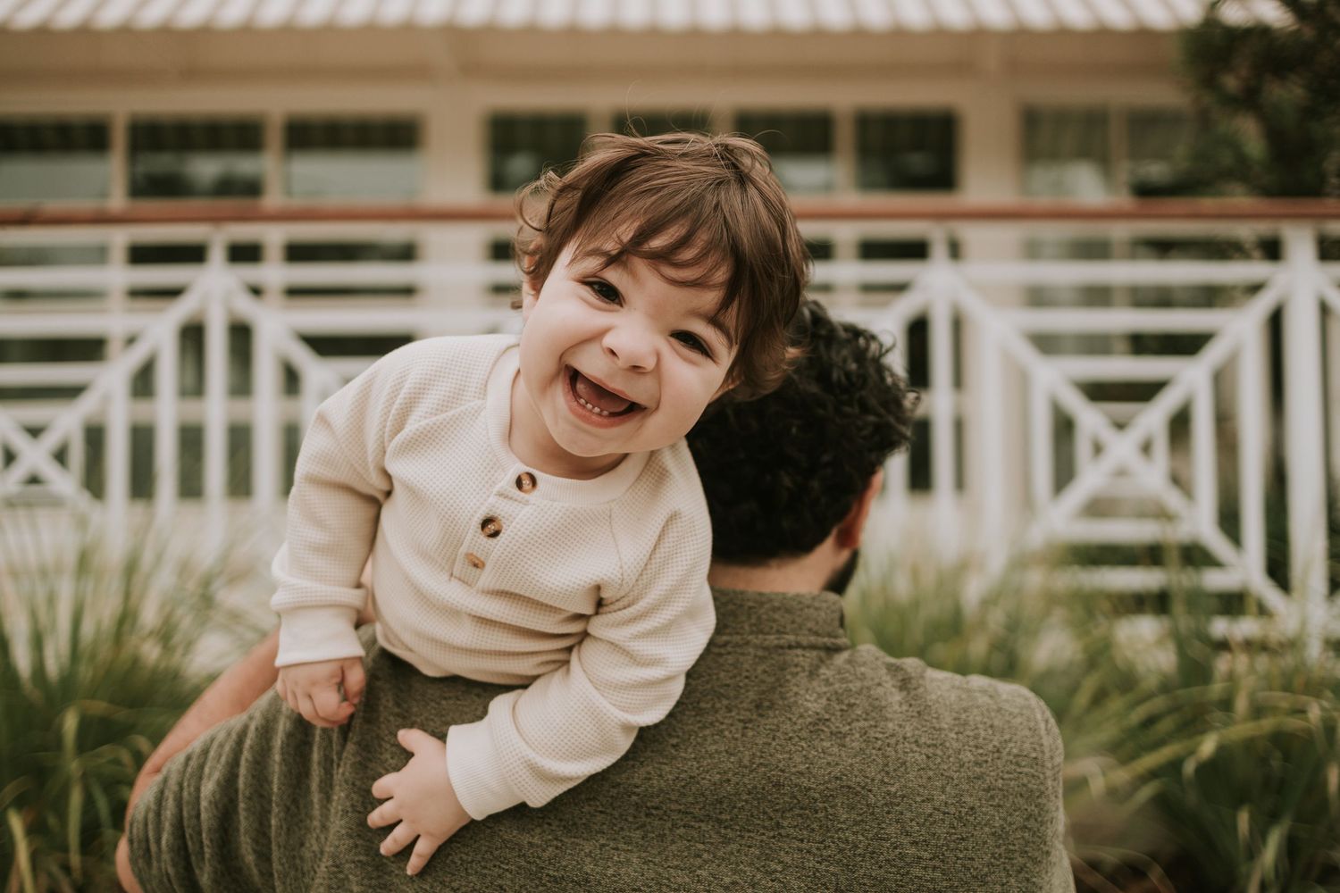 A joyful toddler laughing while being held up by a parent outdoors near a white fence and green plants.