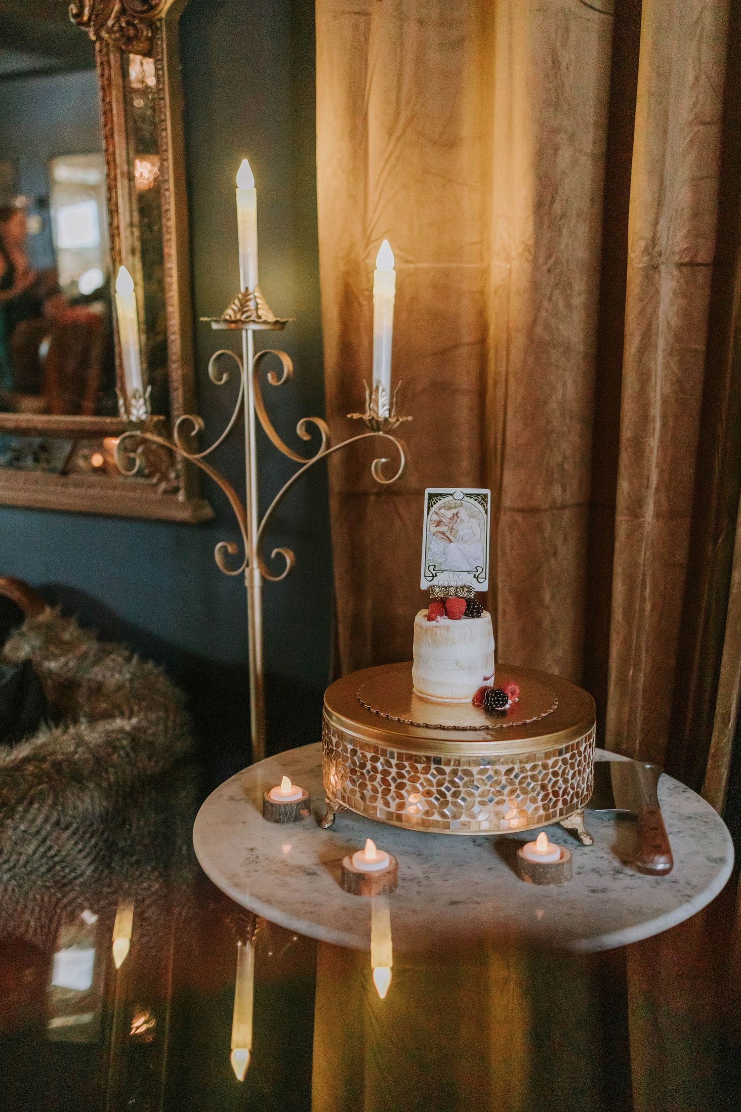 Elegant wedding cake on gold stand with candelabra, taper candles, and mirror in romantic vintage venue setting.