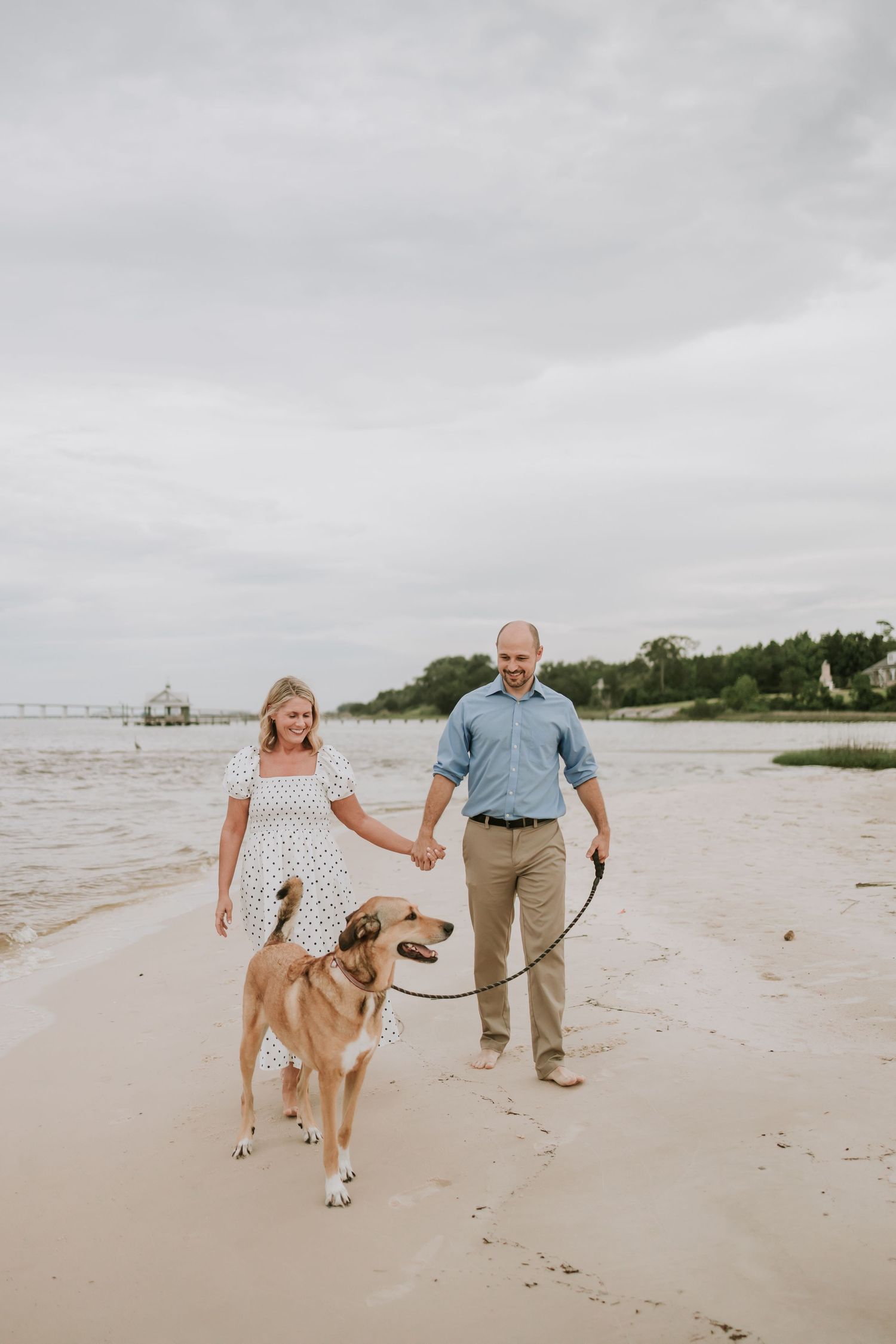 Couple walking a large dog on a sandy beach, woman in white dress, man in blue shirt, overcast sky.