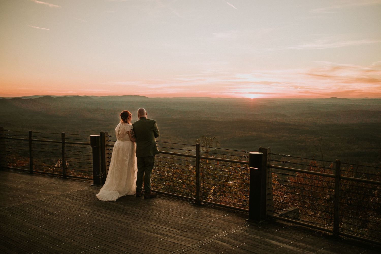 A couple in wedding attire stands at an observation deck overlooking a vast landscape during sunset.