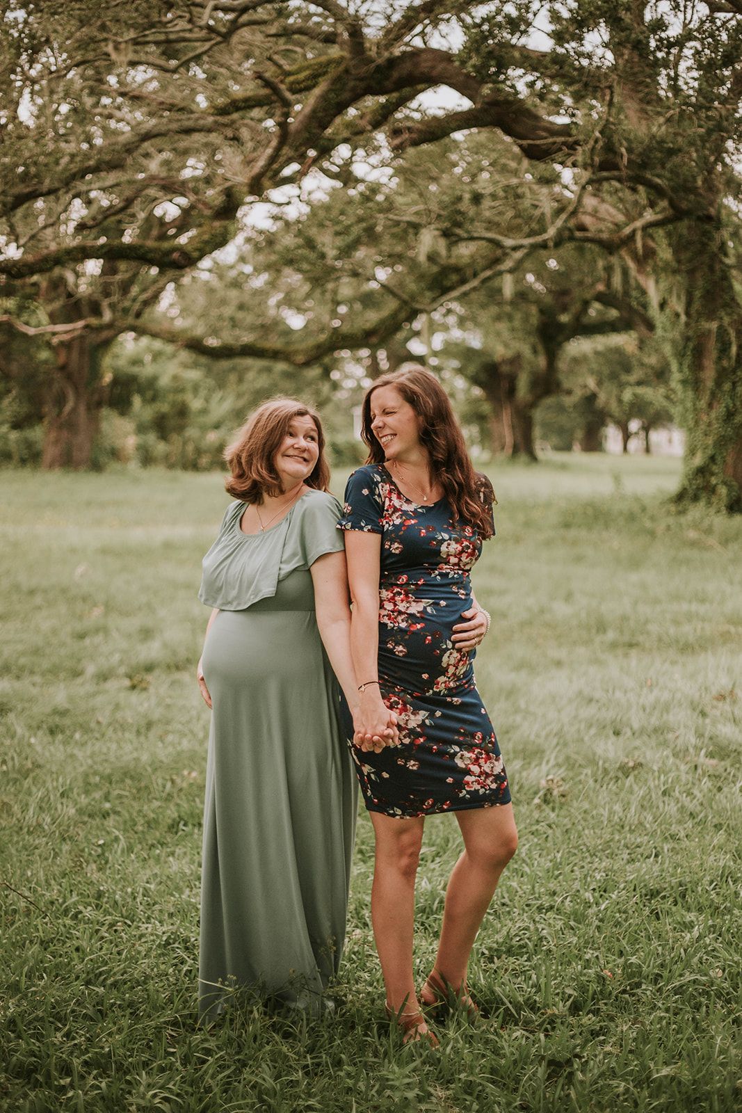 Two expectant mothers in floral dresses share a joyful moment under trees in a park.