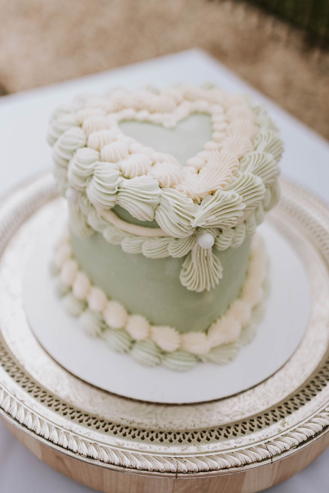 A mint green mini cake decorated with white buttercream piping in a heart pattern and bow design sits on an ornate silver platter.