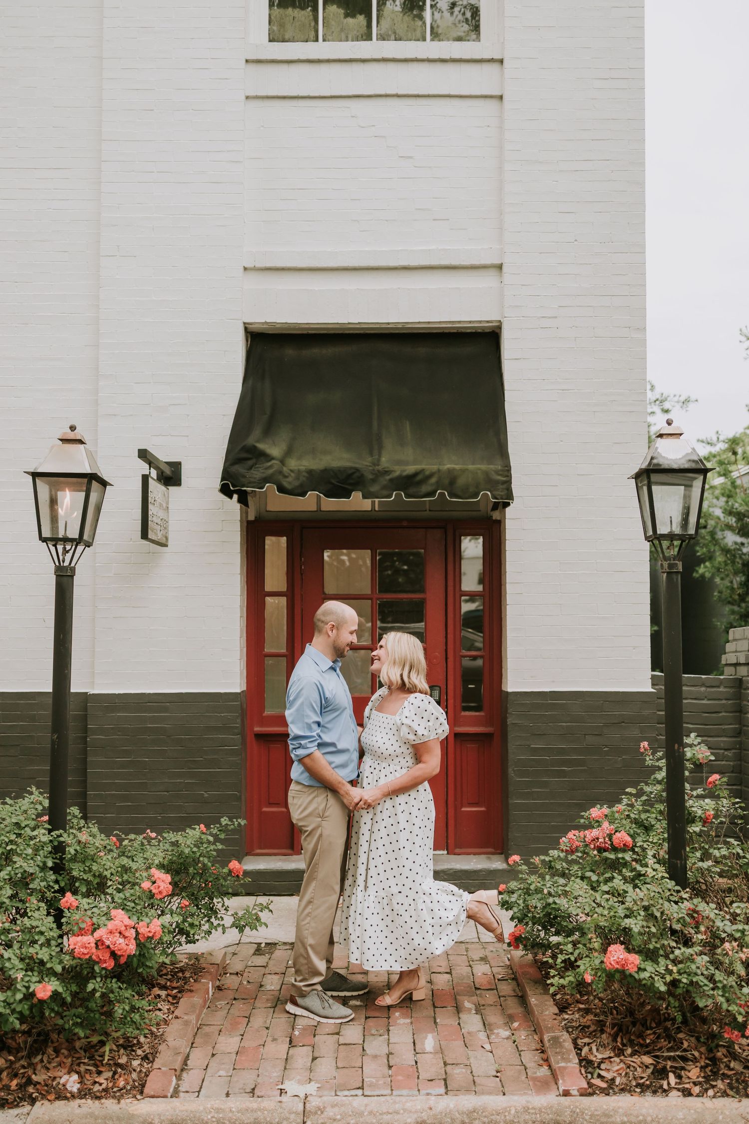 Couple embracing in front of a red door on a brick path, surrounded by pink roses and gas lanterns.