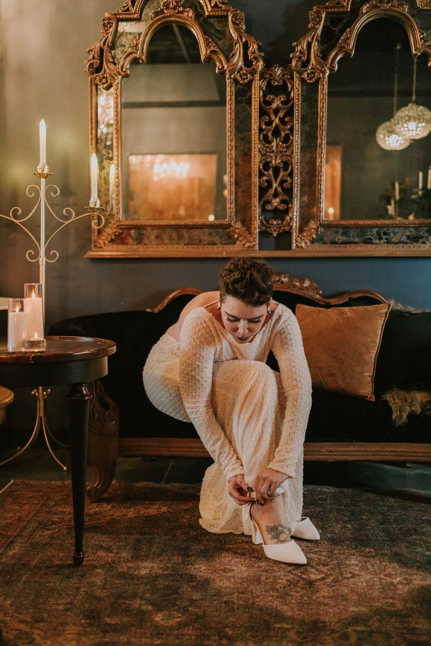 Bride in white lace gown adjusting her heels on ornate rug beside vintage sofa with gold mirror backdrop.
