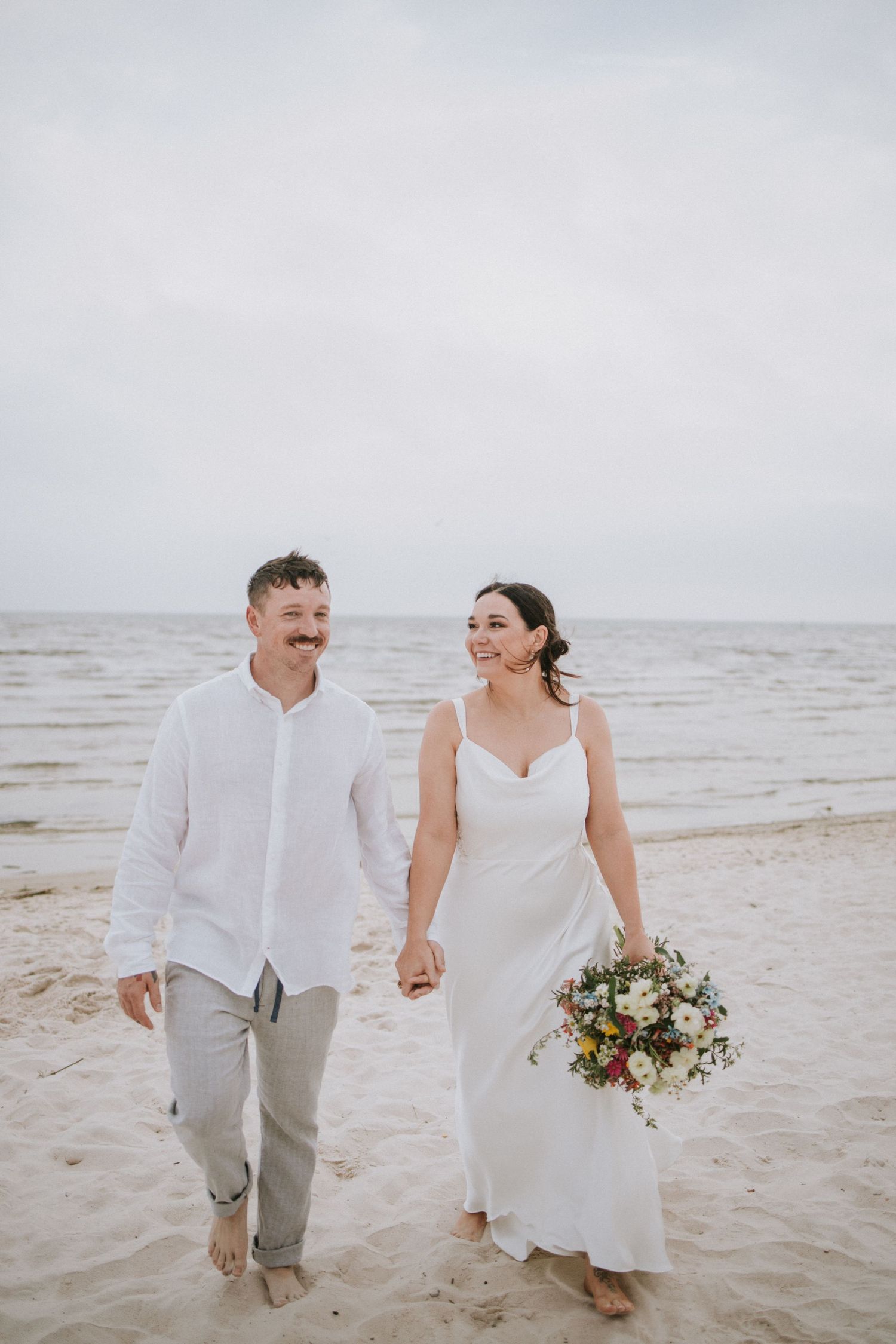 Happy couple walks hand-in-hand on beach, bride in white gown with bouquet, groom in white shirt and gray pants.