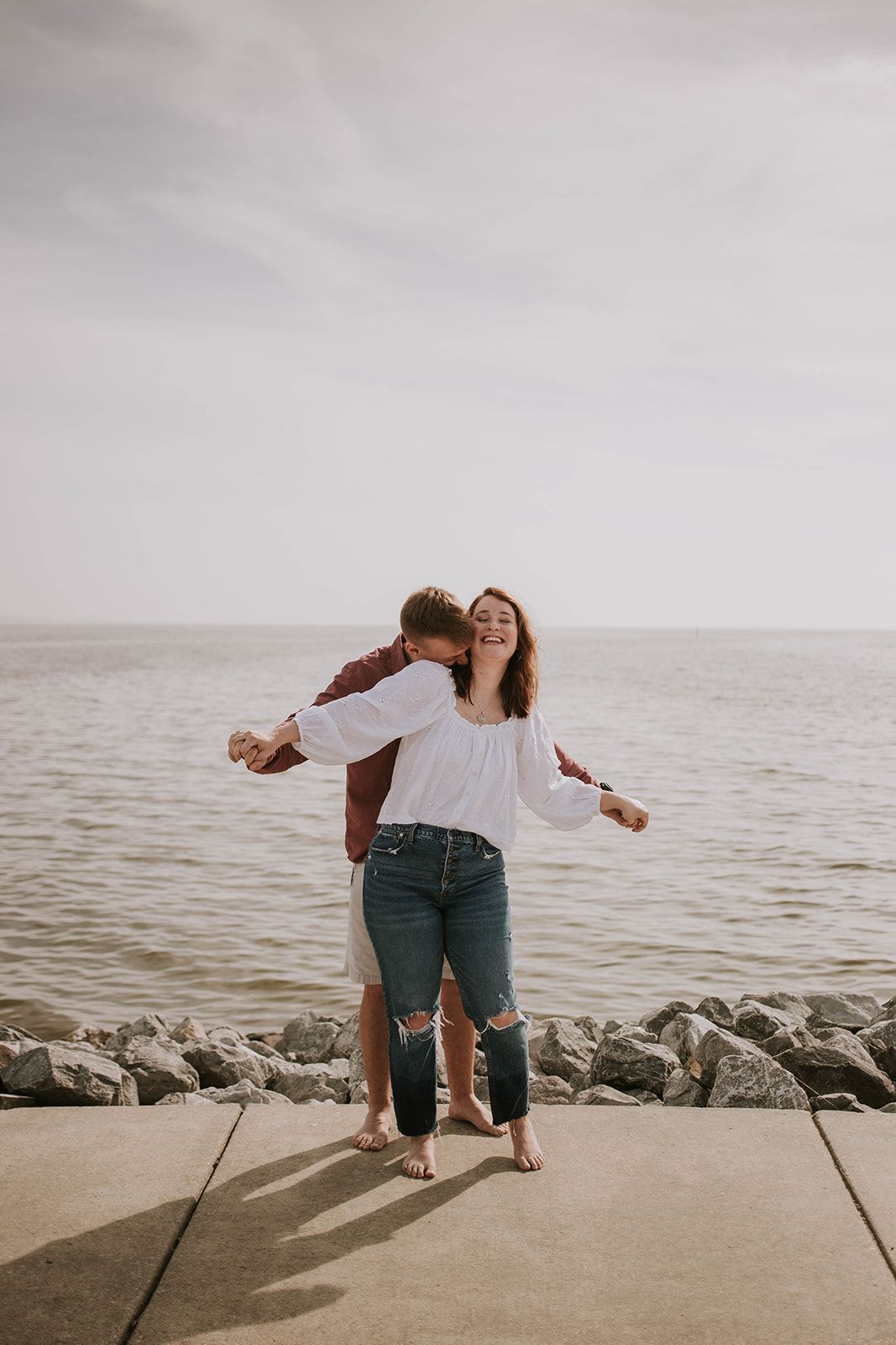 Playful couple embracing on a rocky pier overlooking a misty ocean.