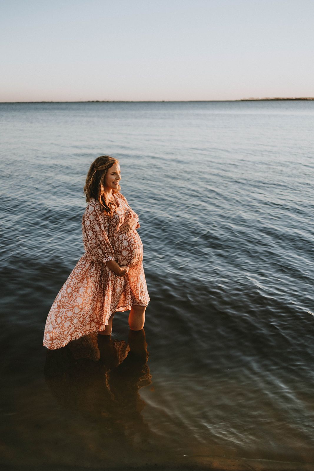 A silhouette stands in flowing dress at the water's edge during sunset.