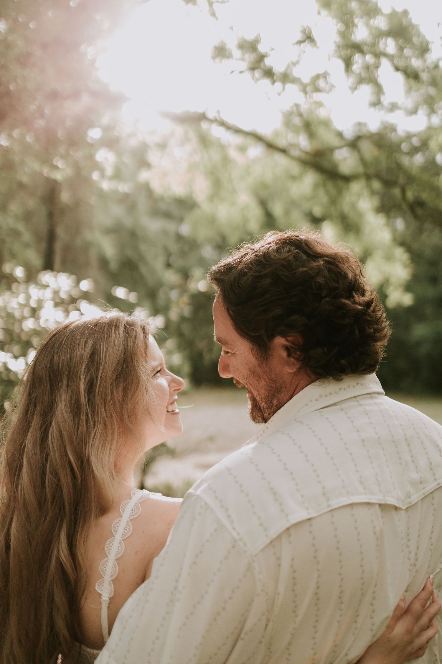 A smiling couple embraces outdoors in soft sunlight, surrounded by lush green trees in a romantic garden setting.