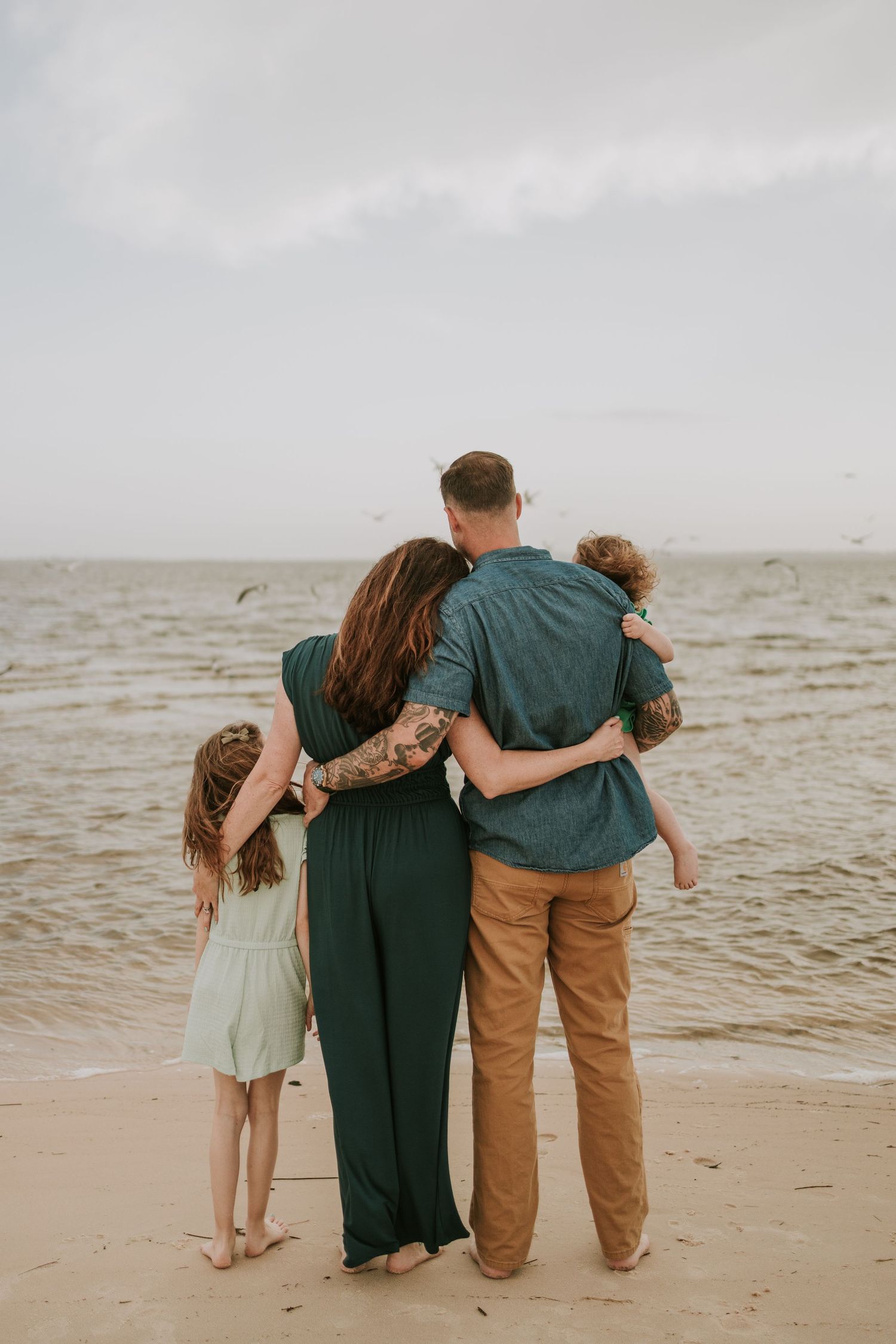 Family of three embraces while standing on a beach looking at ocean on cloudy day.