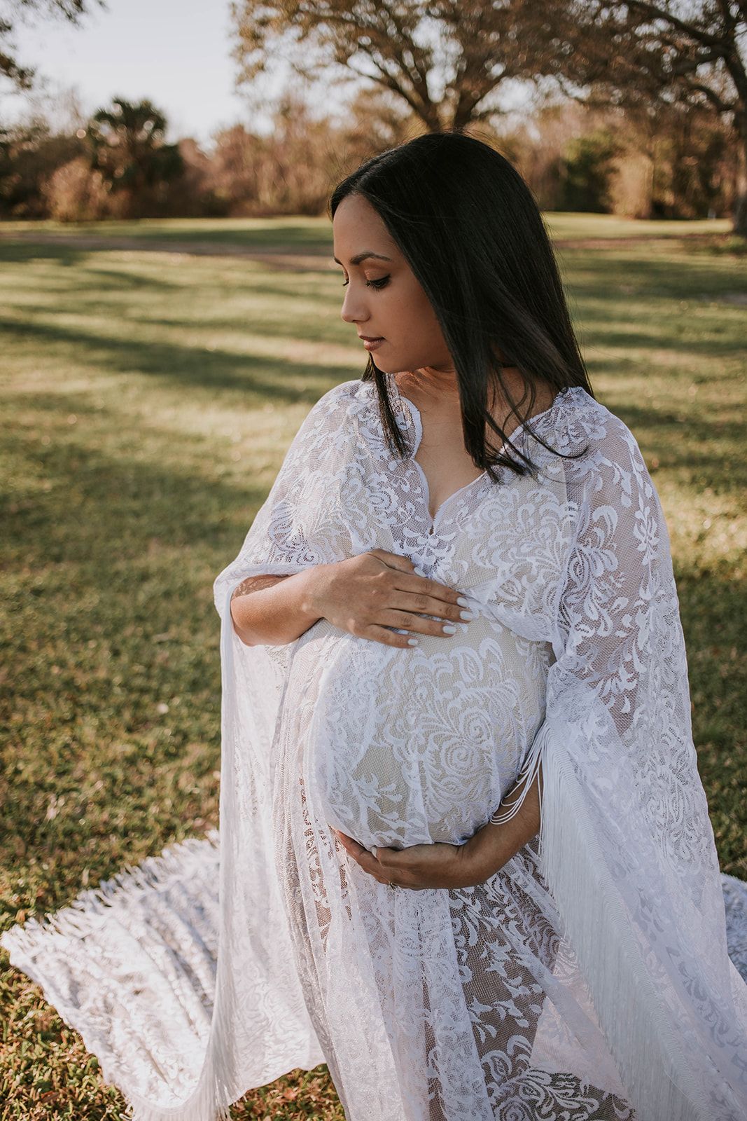 Expectant mother in white lace dress gently touches her belly while standing in a sunlit garden setting.
