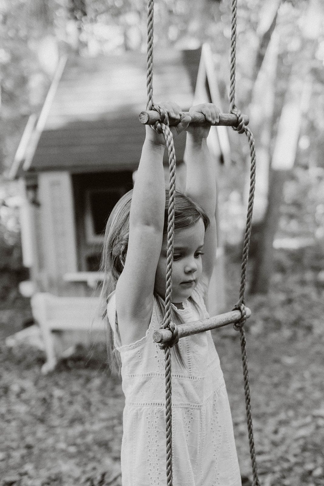 Child in a white dress playing on a rope swing in a black and white photograph.