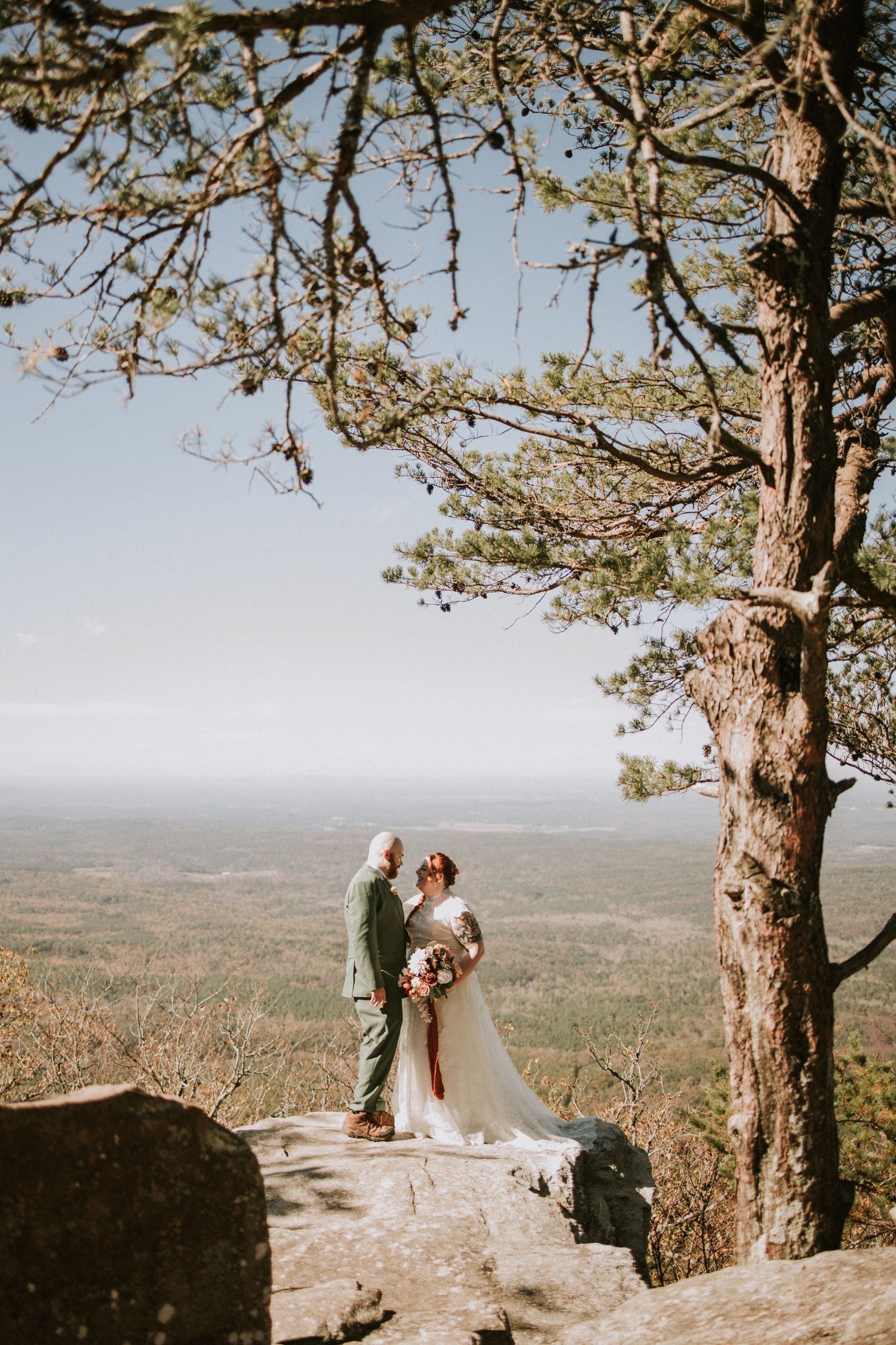 Two people share an intimate moment on a rocky mountain overlook with a vast landscape stretching into the distance.