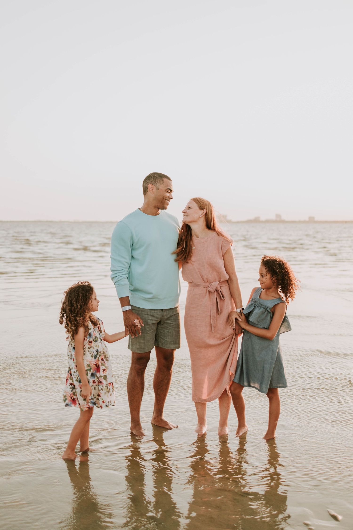 Family of four standing barefoot in shallow beach water, parents and two young girls in pastel outfits smiling.