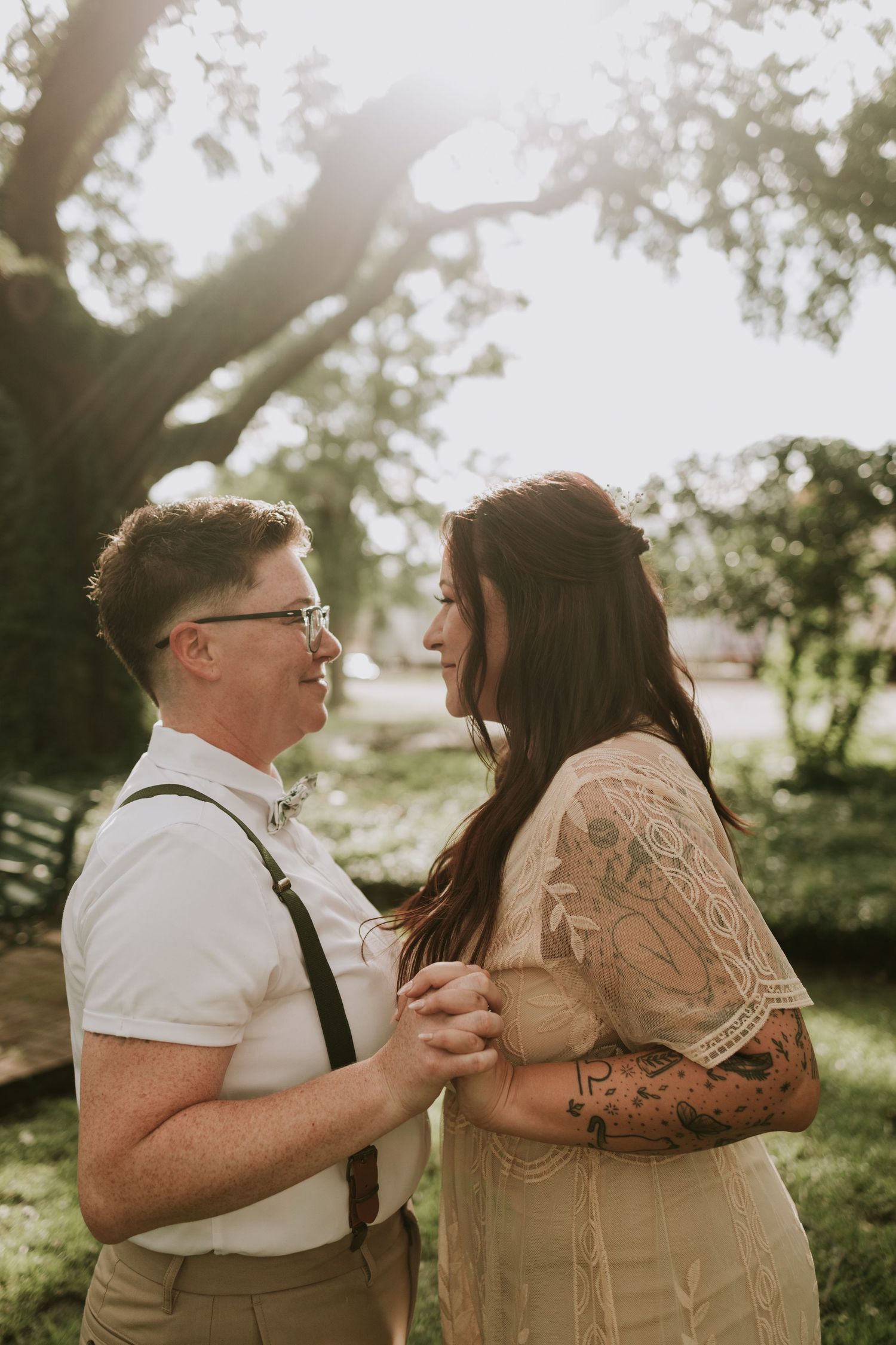 Couple sharing a romantic moment outdoors, woman with tattoos in lace dress, man in suspenders, golden sunlight filtering through trees.