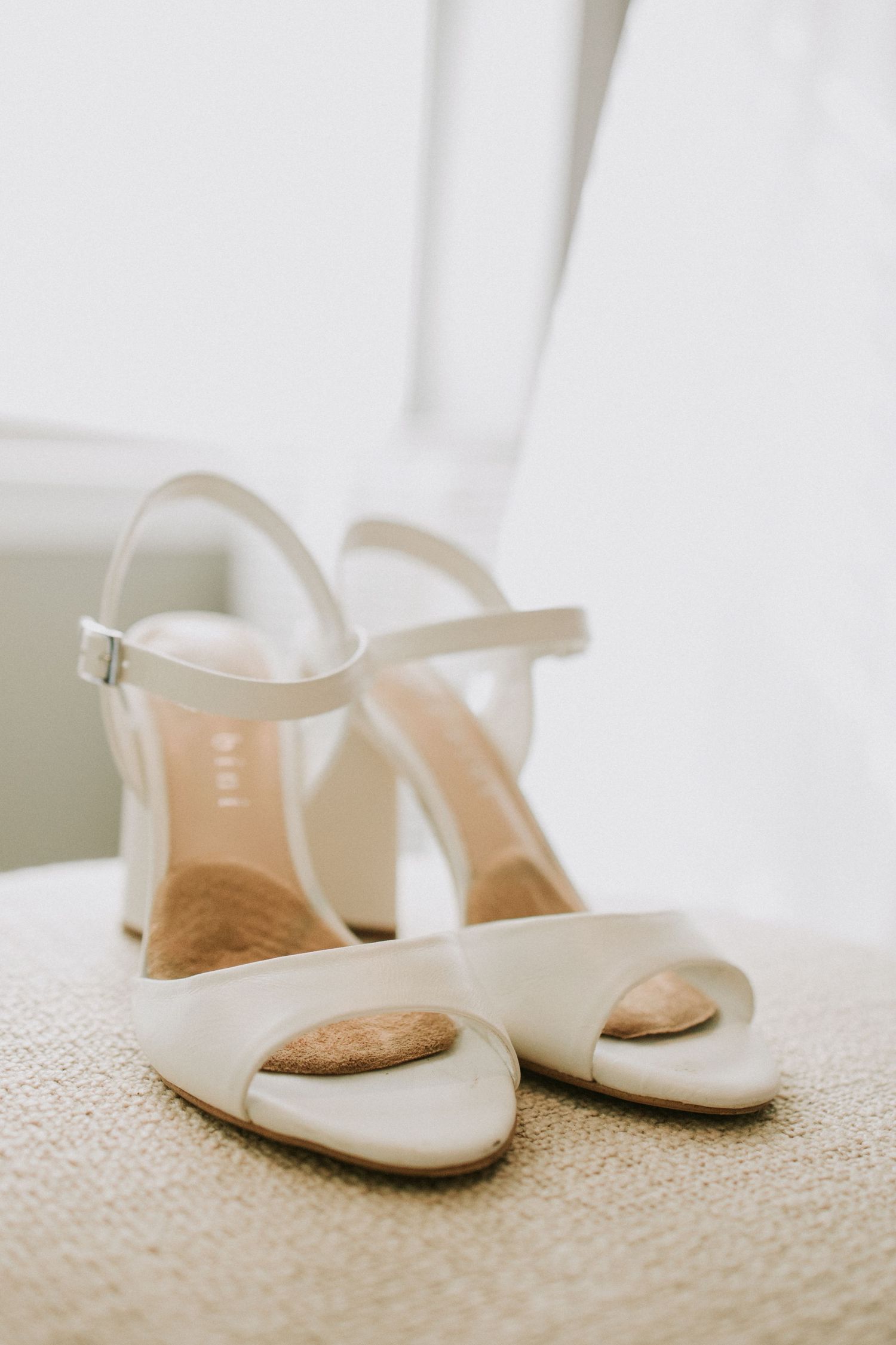 Elegant white bridal heeled sandals with ankle straps displayed on a beige textured surface near a window.