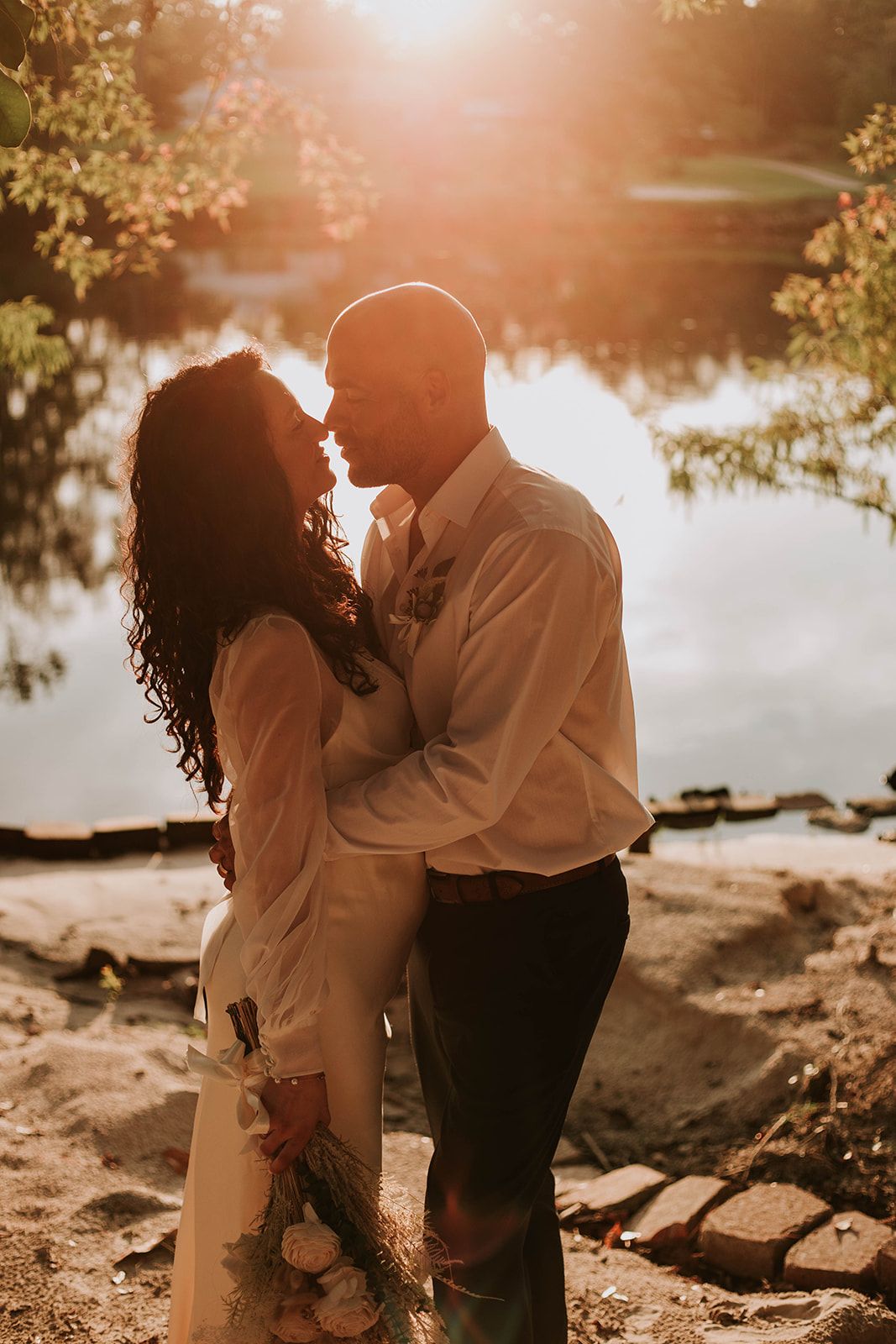 A romantic couple embraces by a serene lake at sunset, creating a dreamy silhouette against the glowing water.