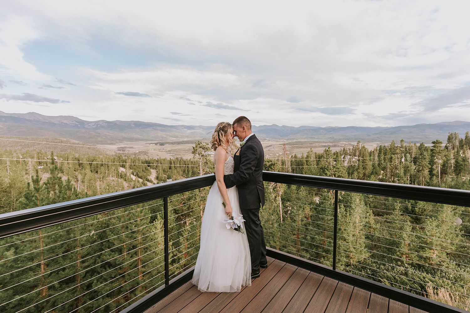 A couple embraces on a wooden deck overlooking a scenic mountain landscape with evergreen trees.
