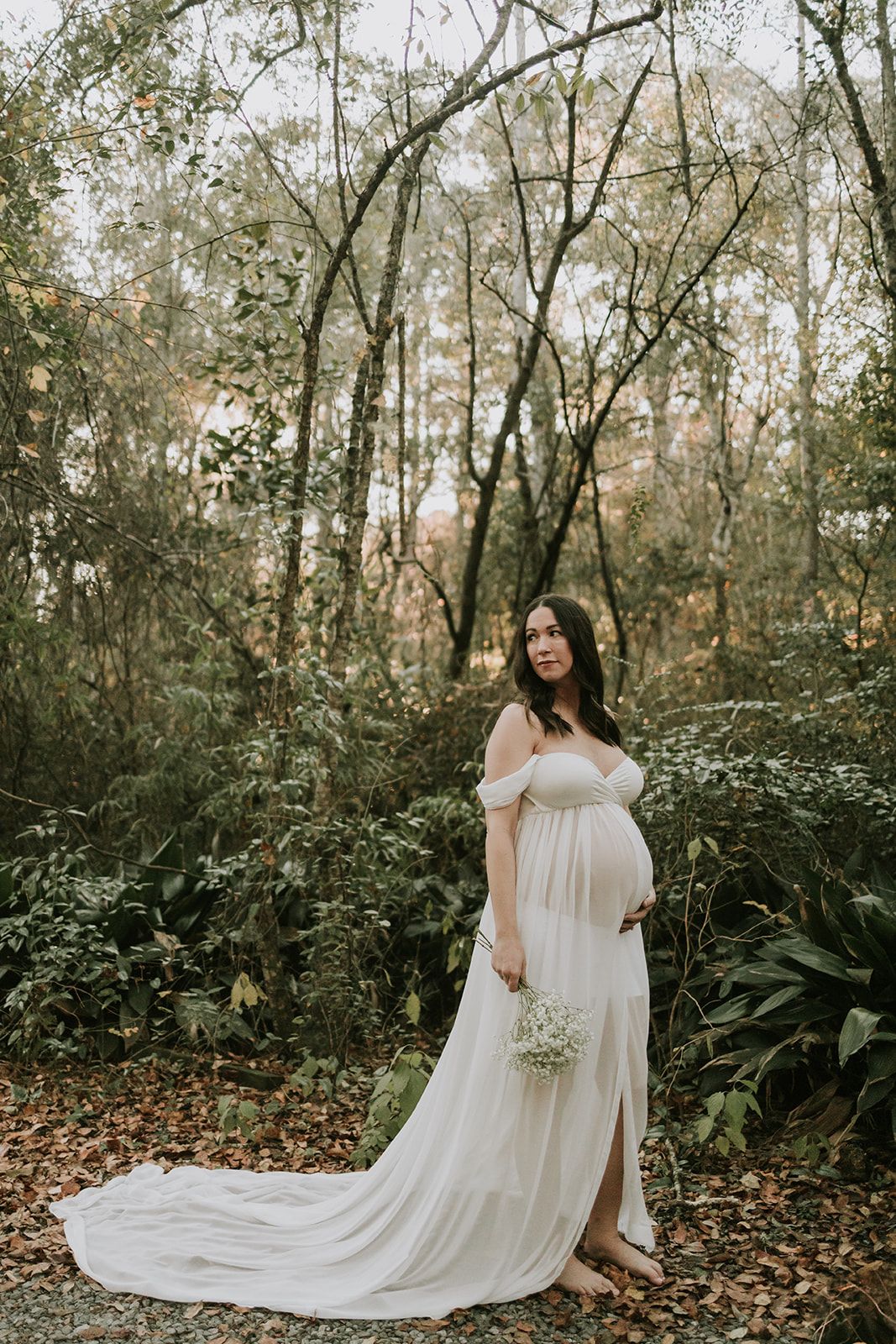 Ethereal maternity photo in white flowing dress among forest trees and fallen leaves.