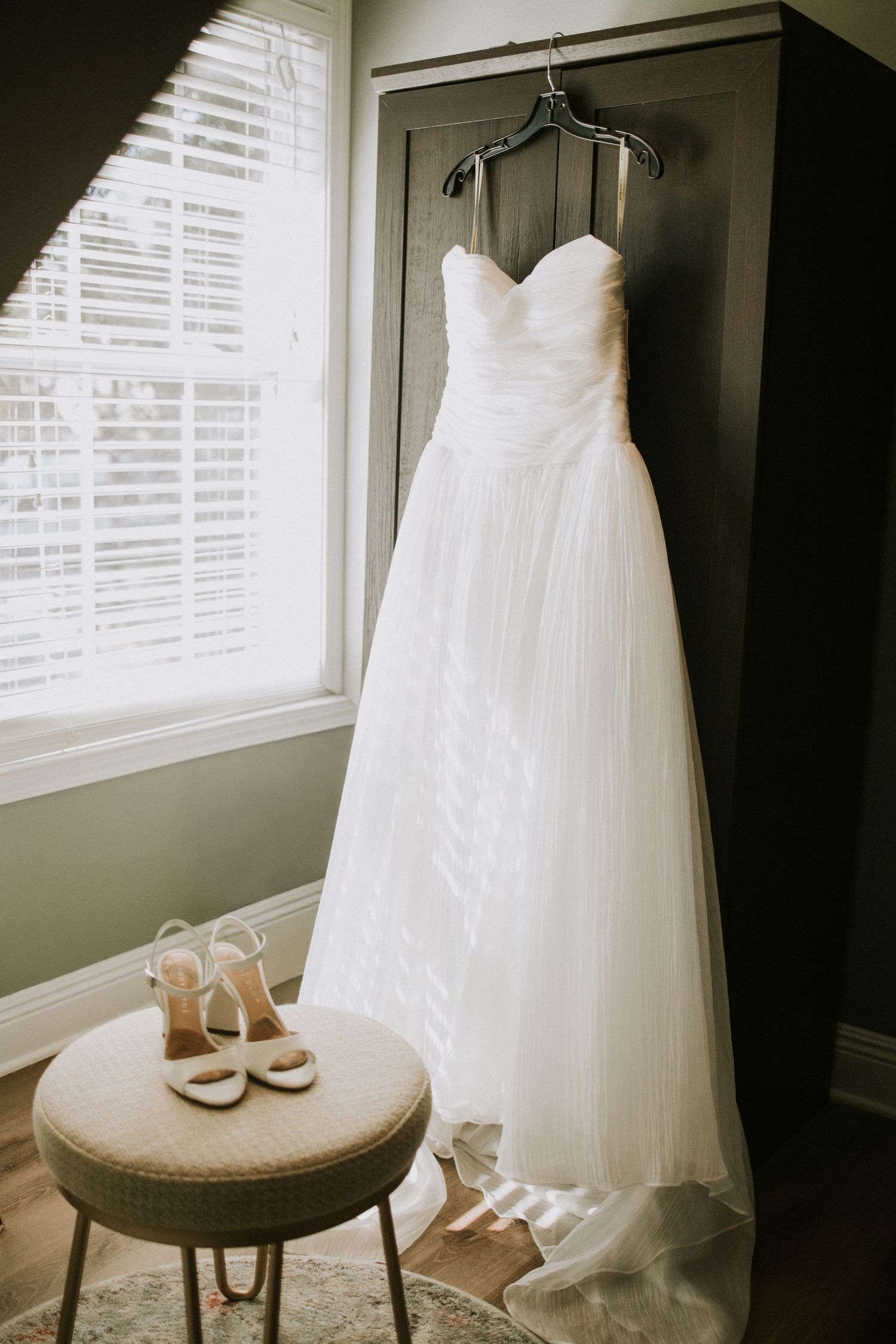 White strapless wedding gown hanging on dark armoire beside window with bridal heels displayed on tufted ottoman.