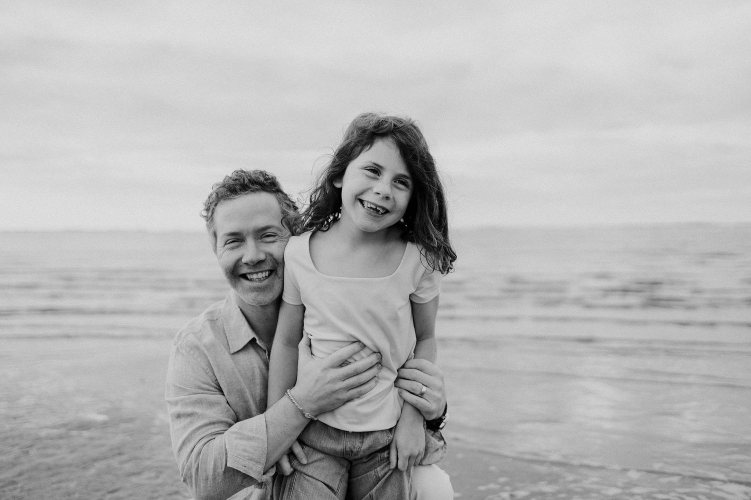Black and white photo of a laughing father and daughter posing together on a beach.