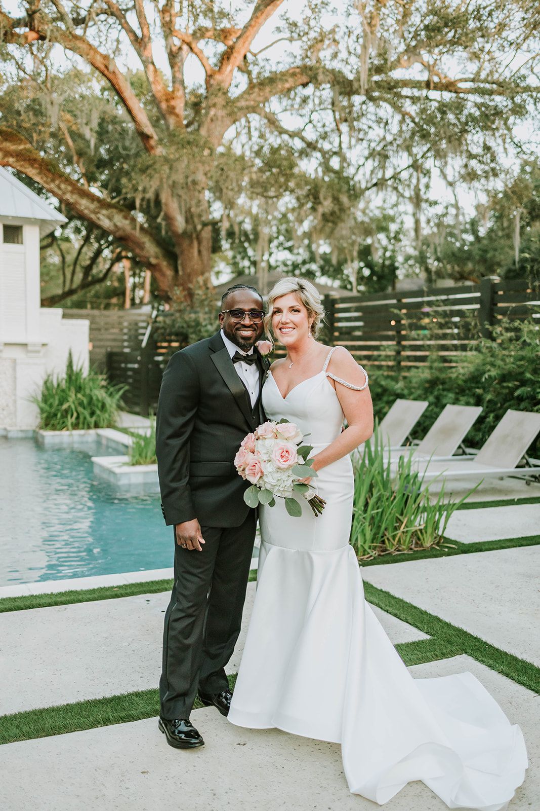 A bride and groom stand together by a swimming pool surrounded by loungers and landscaping in an outdoor garden setting.