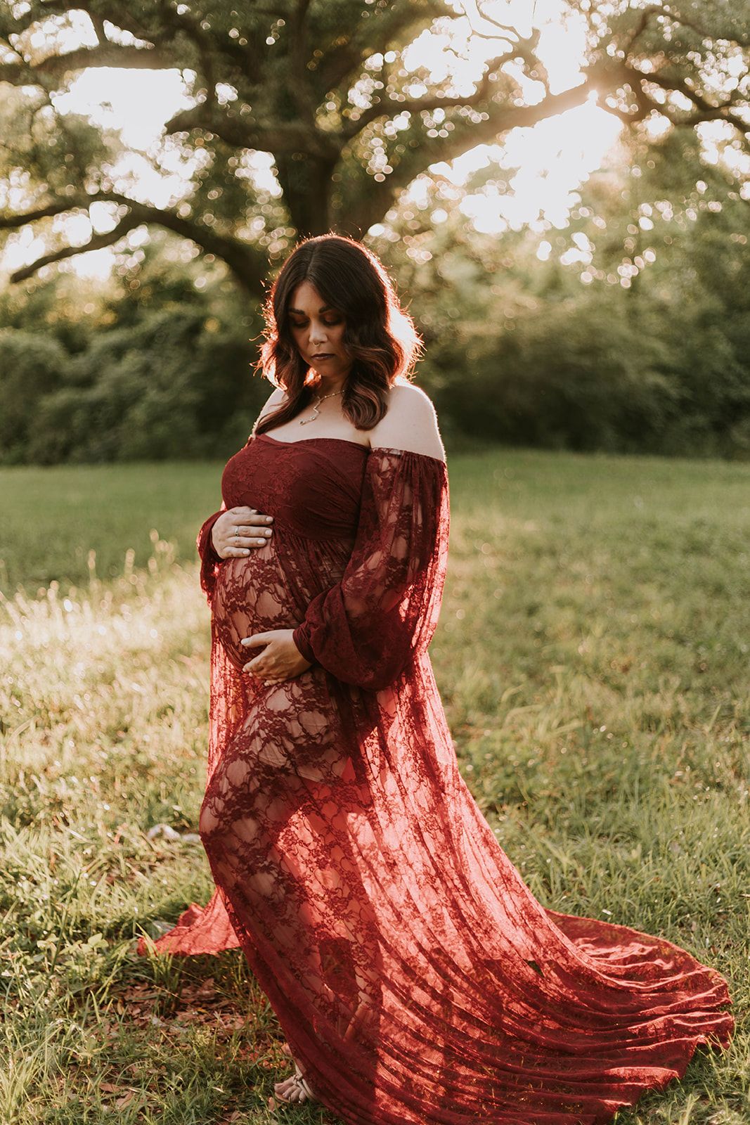 Person in flowing burgundy lace dress poses in sunlit meadow during sunset.