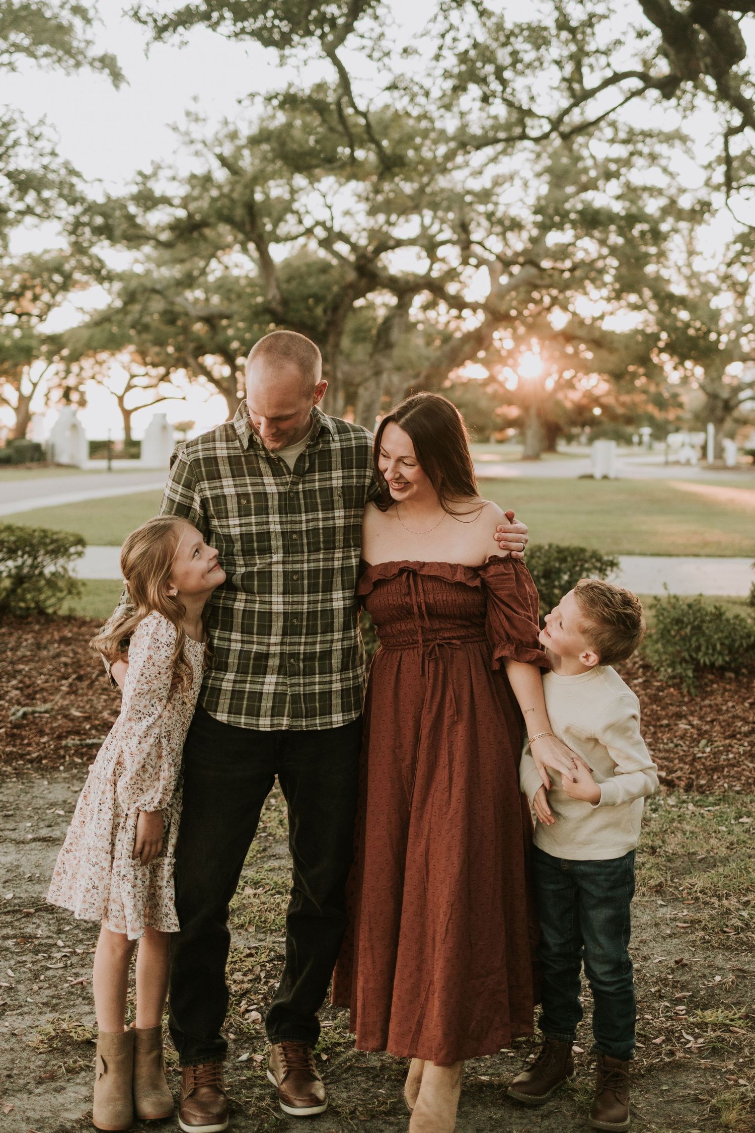 Family of four laughing together outdoors at golden hour, parents with young daughter and son among oak trees.