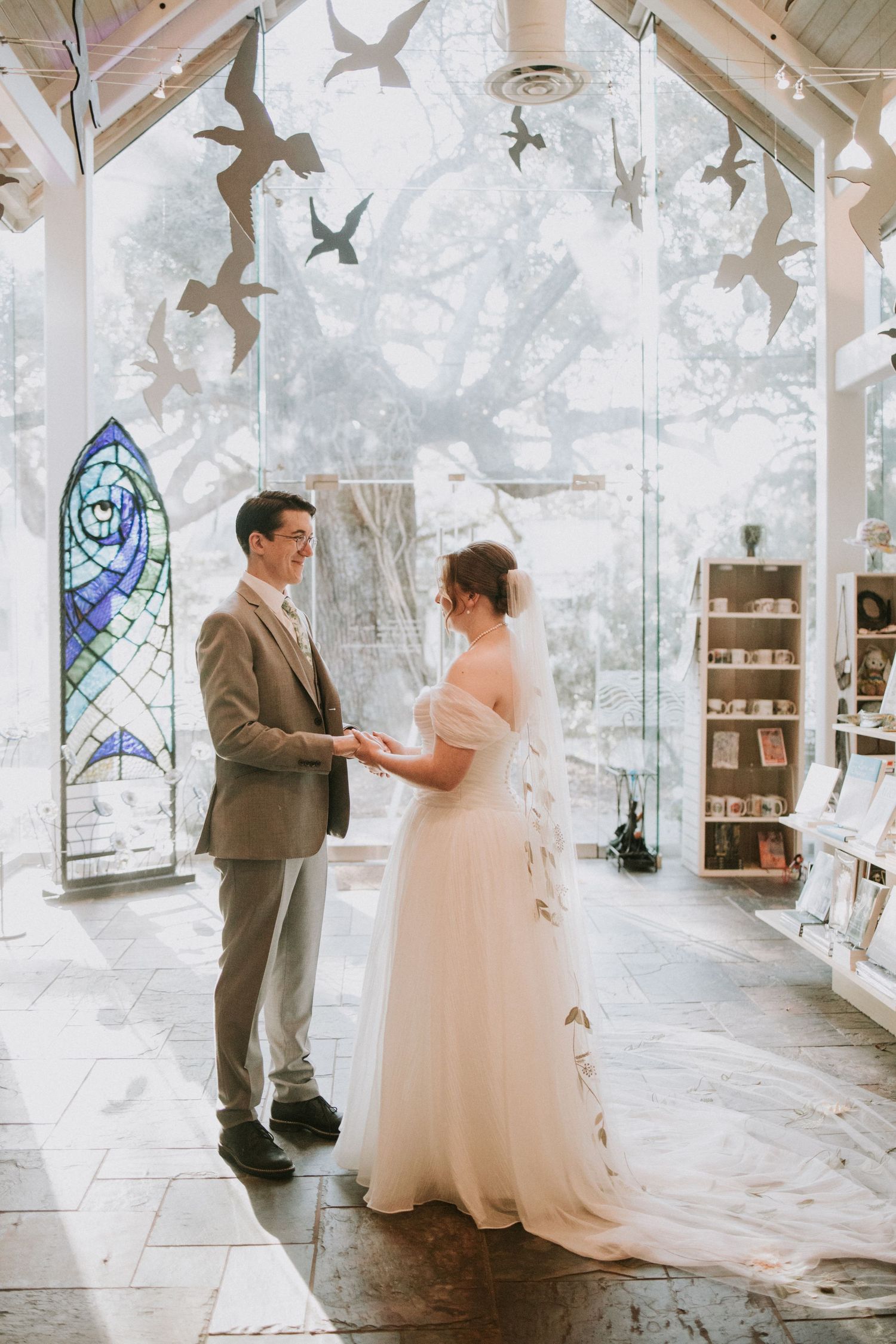 Bride and groom exchange vows in a sunlit chapel with stained glass windows and bird sculptures overhead.