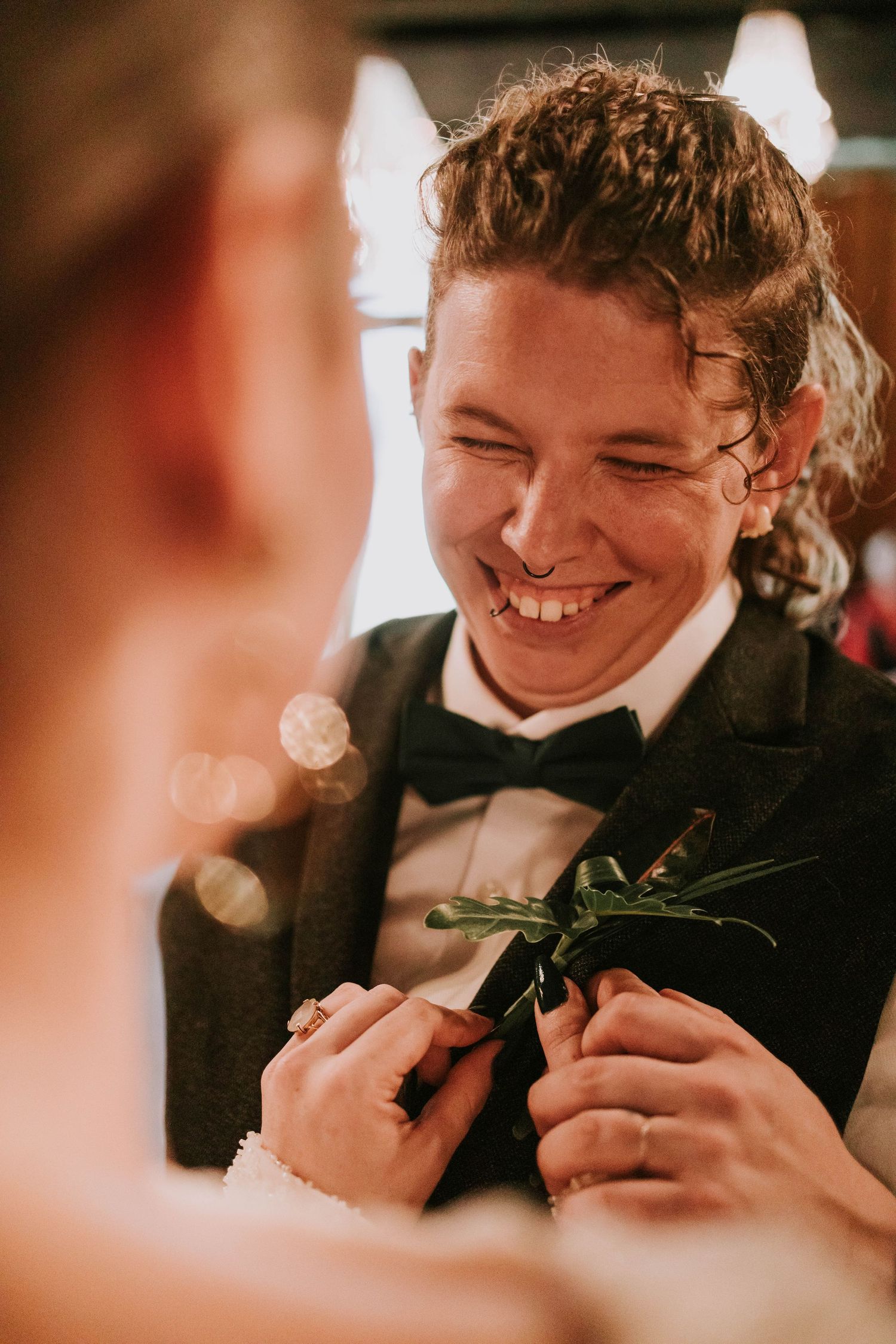 Smiling groom in black tuxedo having boutonniere pinned on lapel at wedding ceremony.