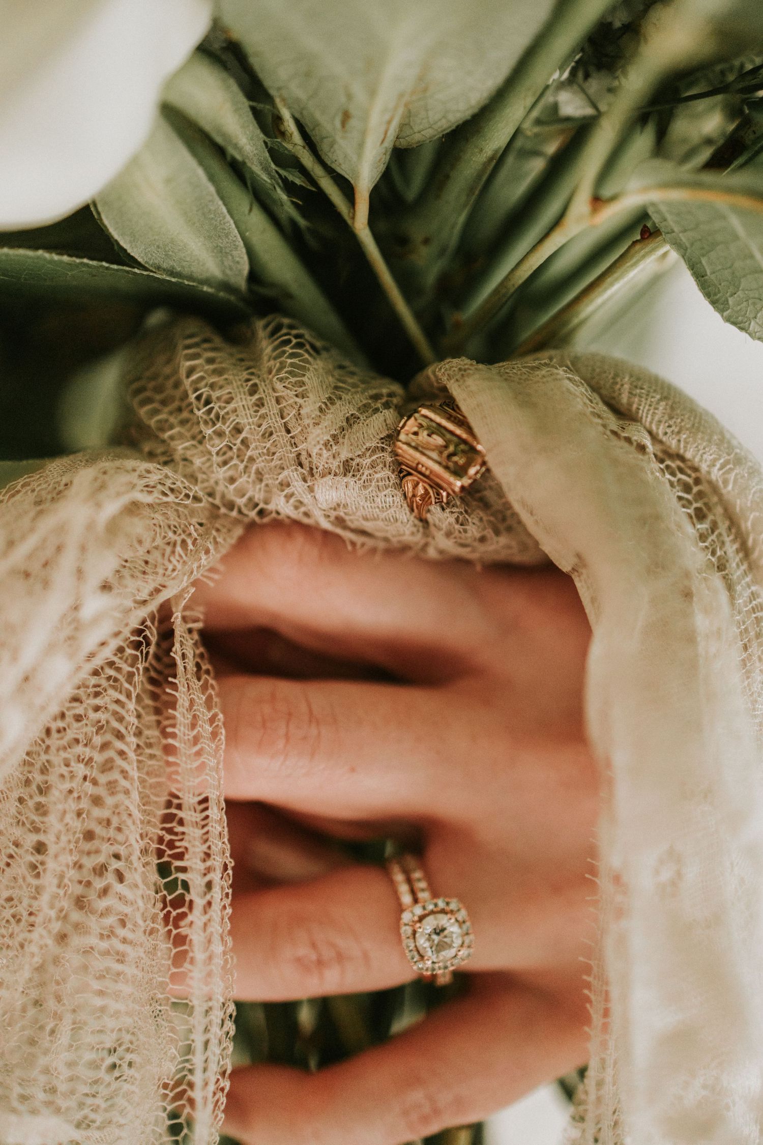 Close-up of a bride's hand holding a bouquet, wearing a halo diamond engagement ring and gold wedding band.
