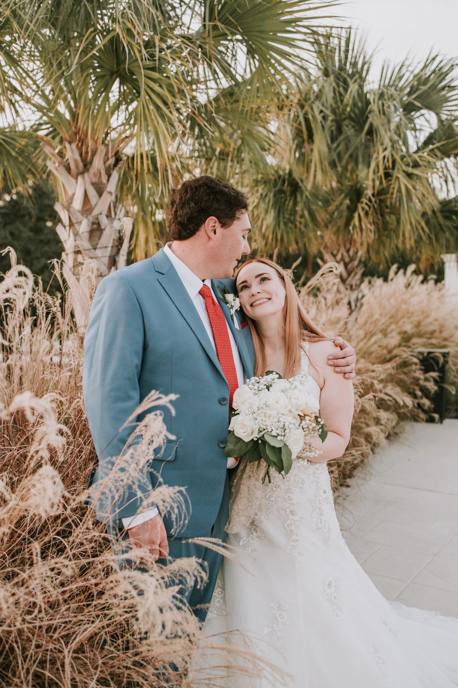 Bride and groom pose outdoors near palm trees, she holds white bouquet, he wears blue suit with red tie.