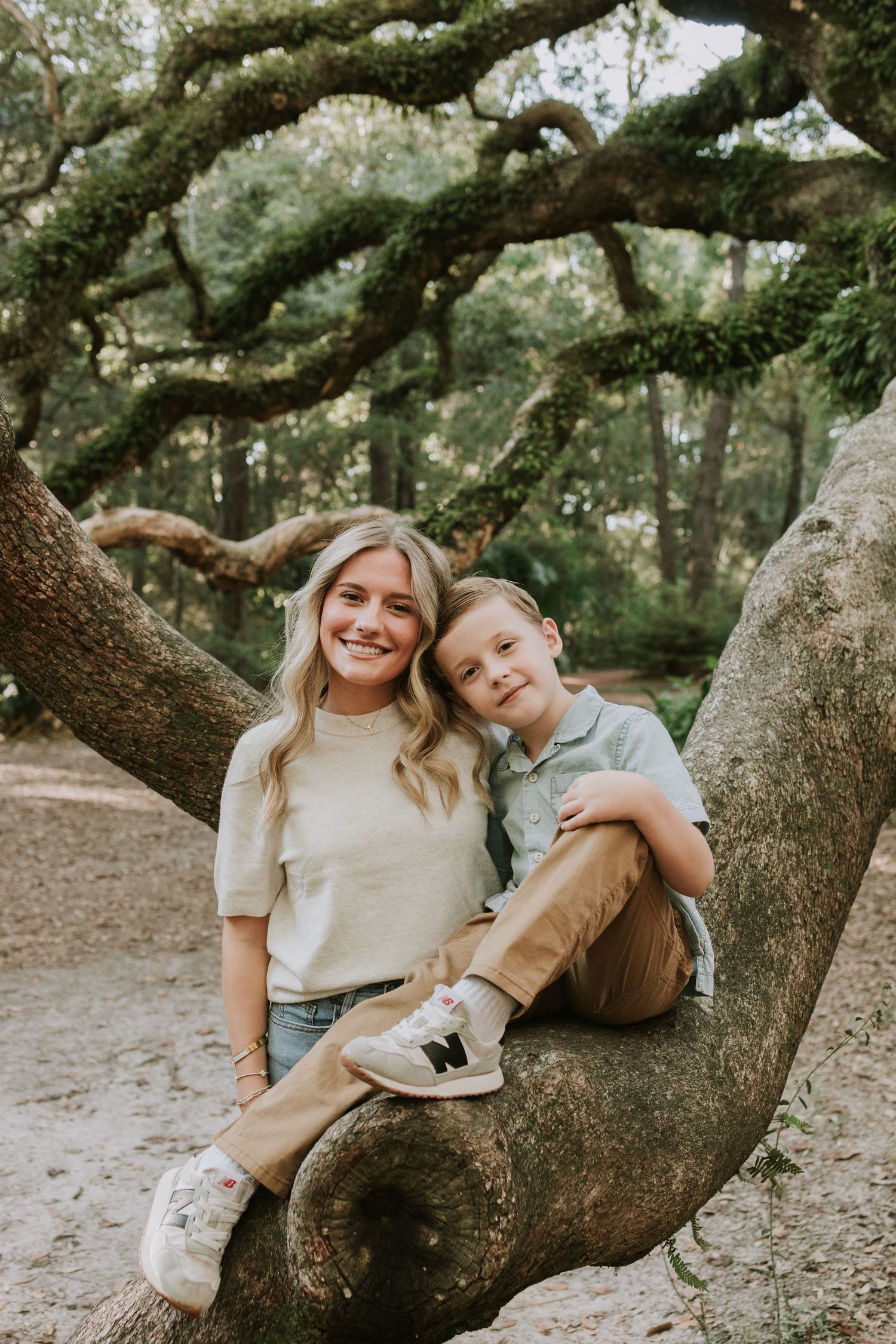 A smiling woman and young boy sit together on a large tree branch in a lush green forest setting.