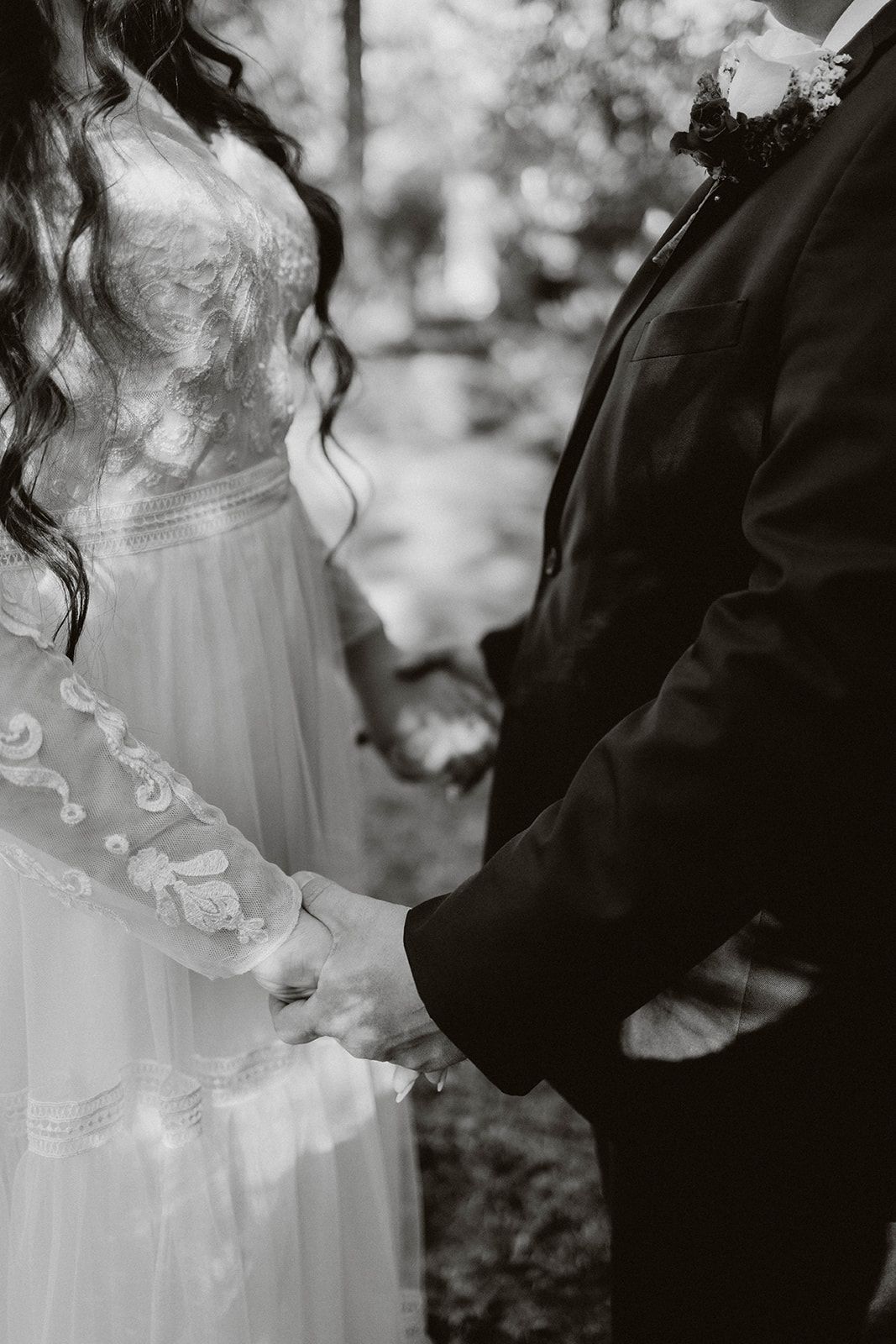 A black and white wedding photo showing couple holding hands, with bride in lace dress and groom in dark suit.