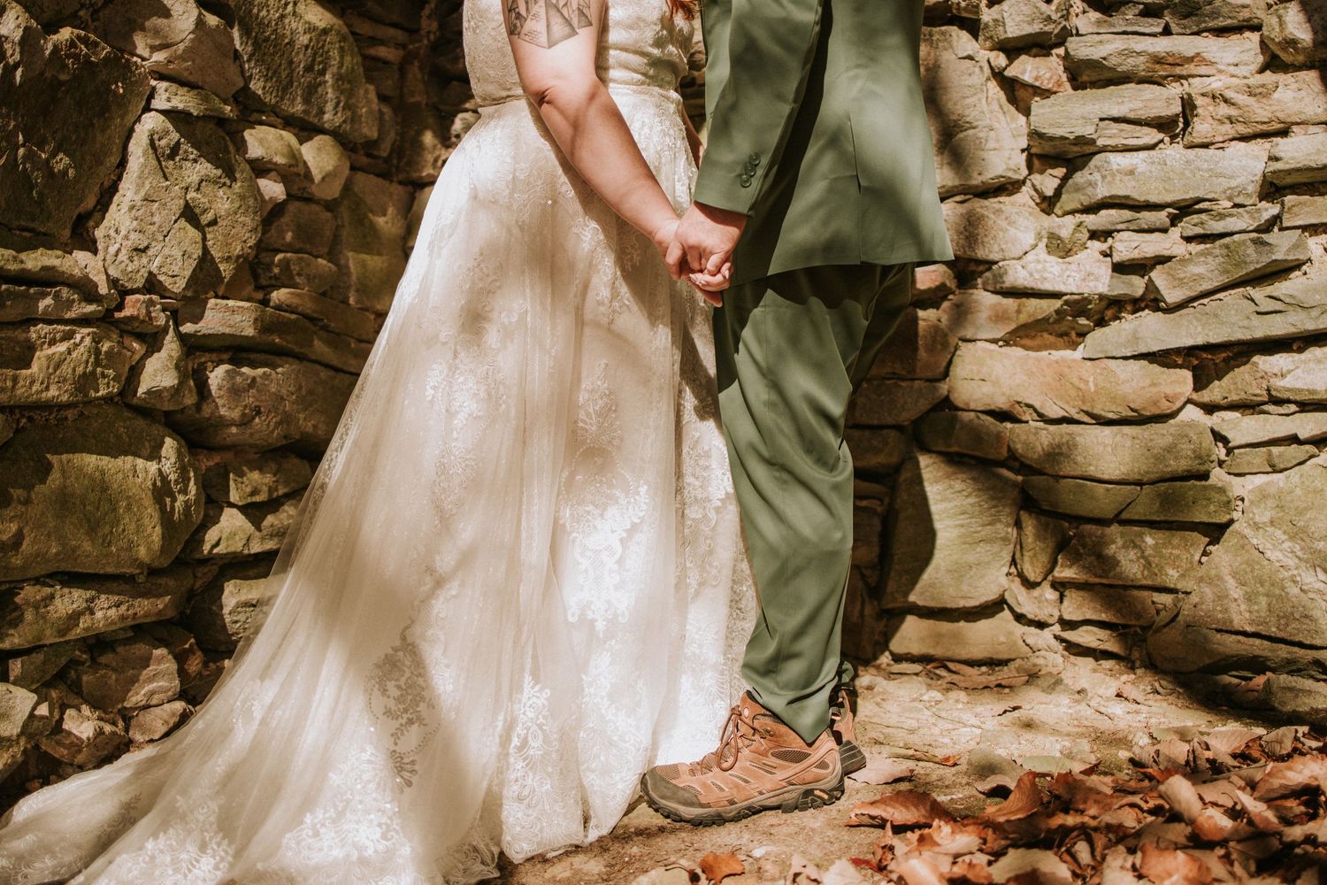 A close-up shot of a bride in a white lace gown and groom in sage green pants against a stone wall.