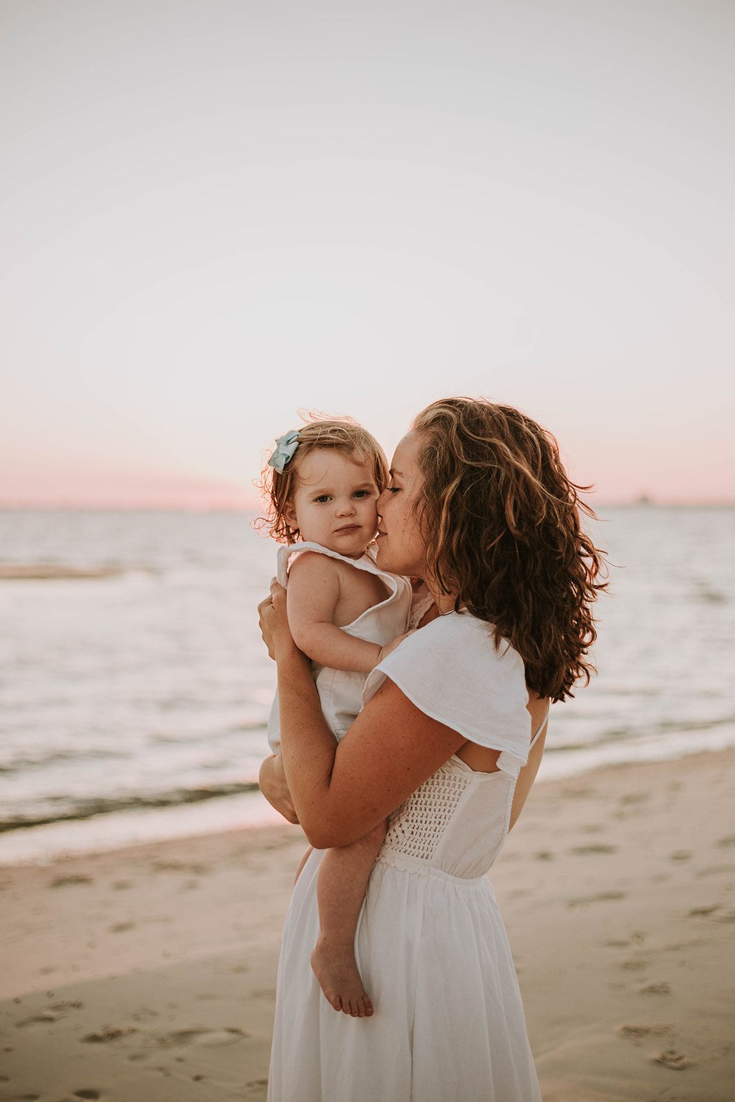 A parent and child share a tender moment at sunset on the beach in white summer dresses.