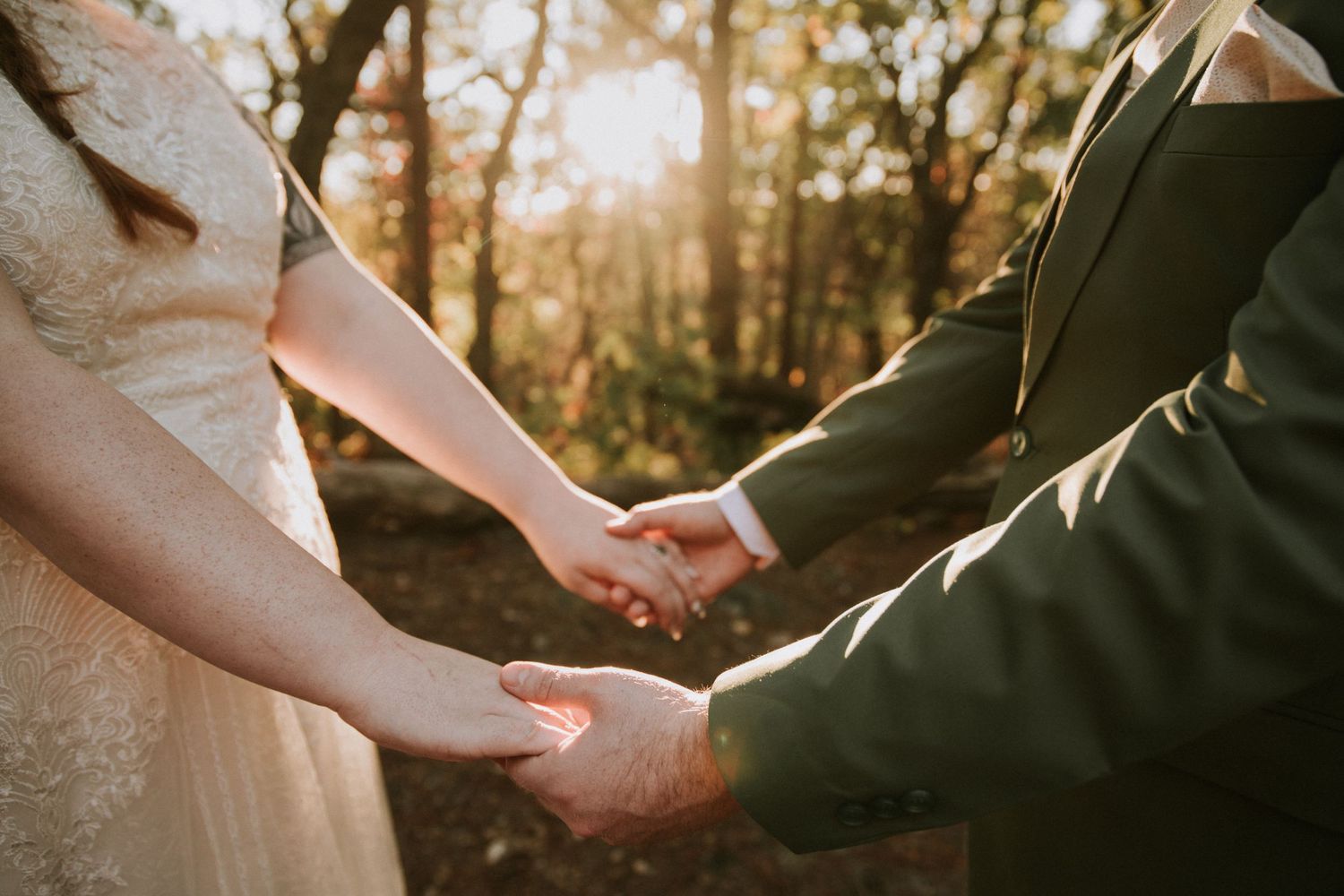 Two people hold hands in golden sunlight filtering through trees while wearing formal wedding attire.