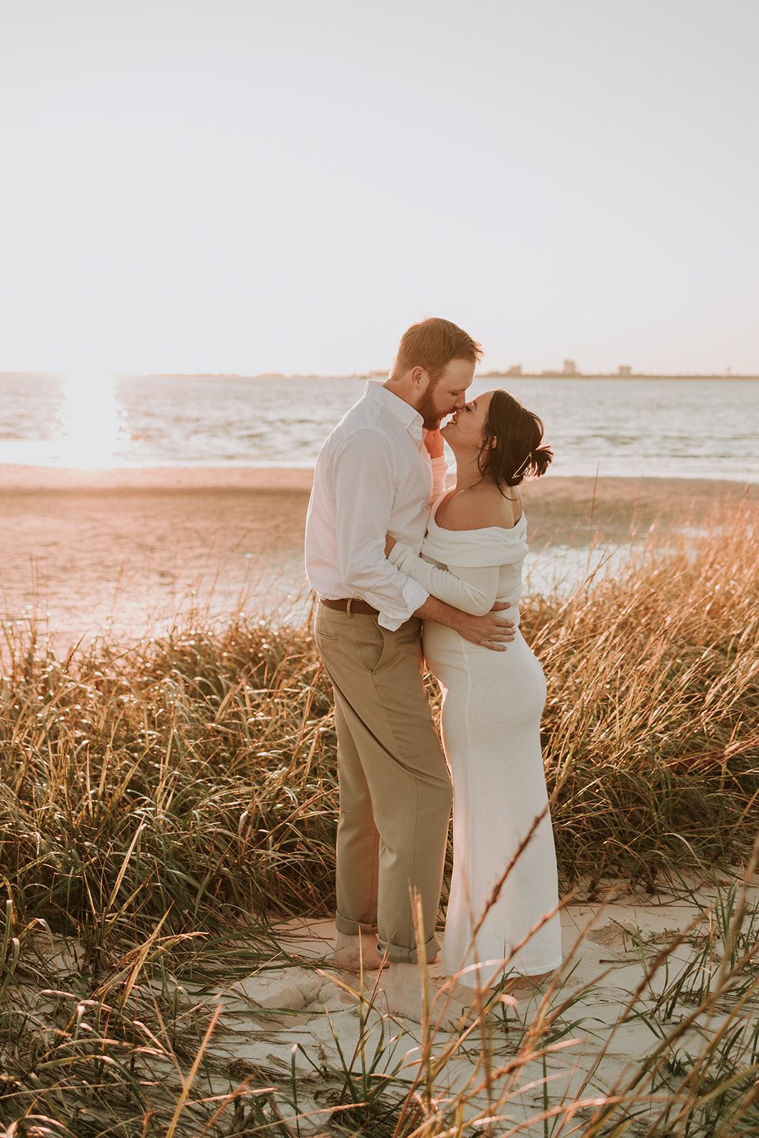 A romantic sunset beach wedding photo shows a couple embracing amid tall grass with the ocean in the background.