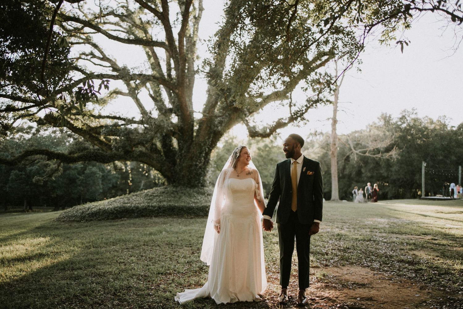 Bride and groom stand beneath a grand oak tree on lush green grounds during their outdoor wedding ceremony.