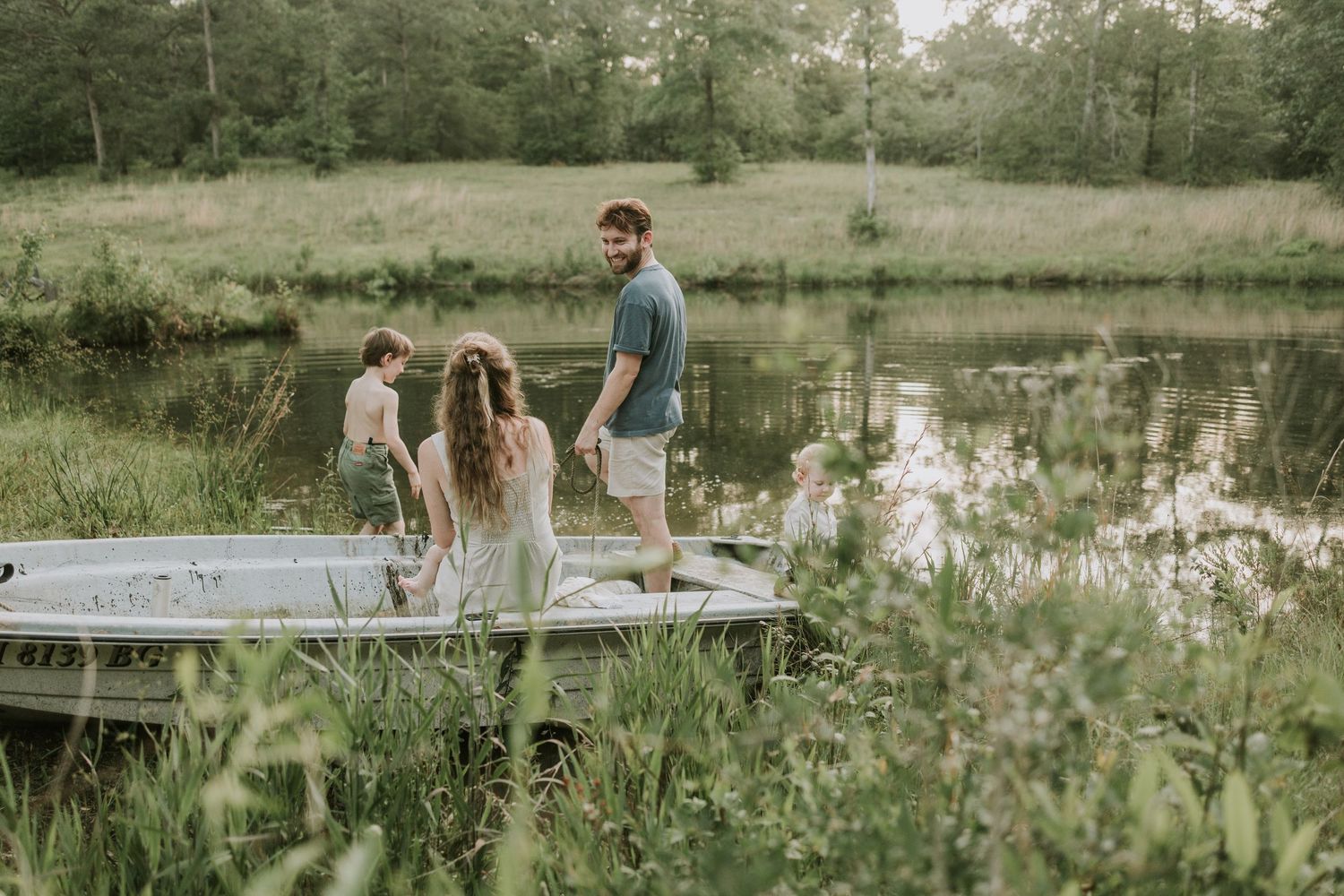 A romantic couple sits by a serene pond surrounded by lush greenery and wildflowers during golden hour.
