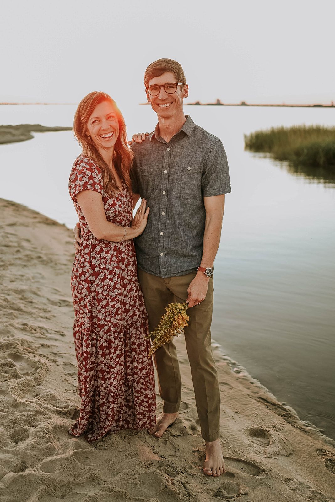 A couple stands together at sunset on a beach near marsh grasses.