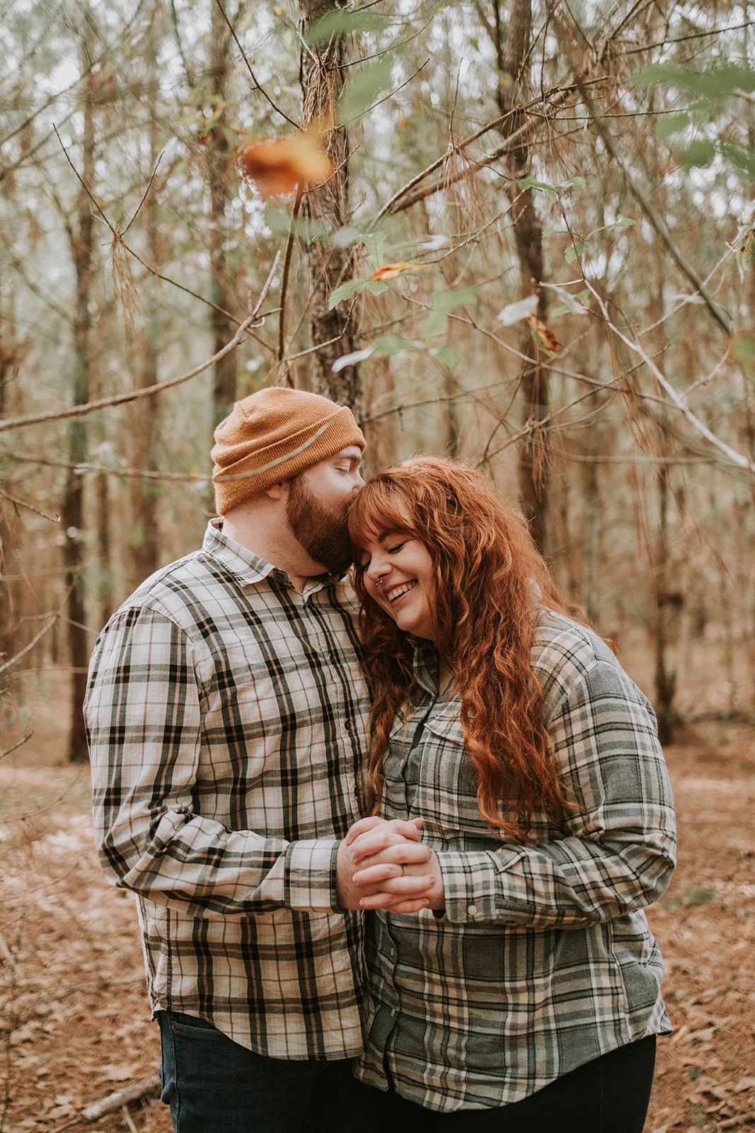 A couple wearing matching plaid flannel shirts embrace while standing together in an autumn forest setting.