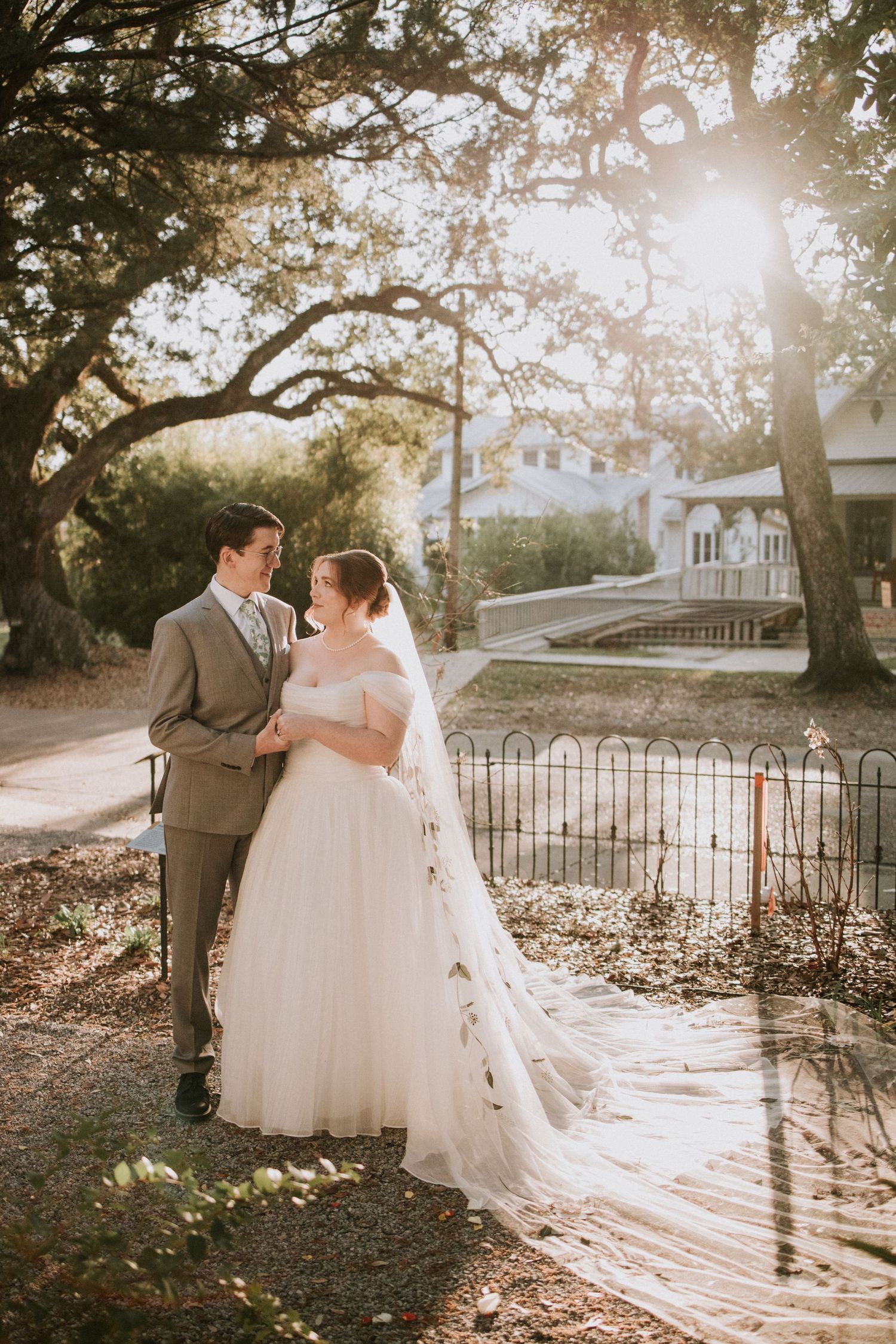 Bride and groom share a romantic moment outdoors under sunlit oak trees, bride in off-shoulder gown with long veil.