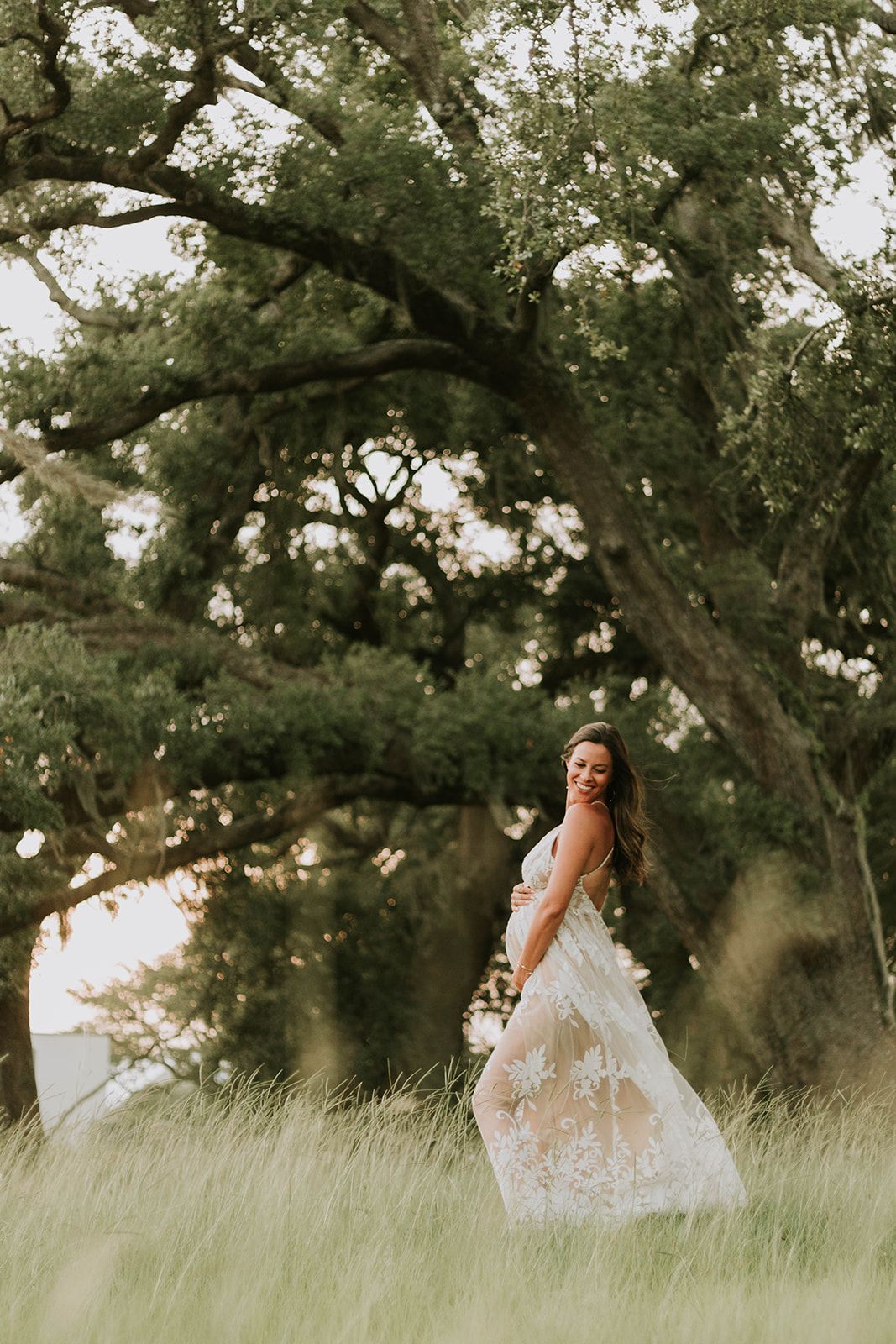 Dreamy maternity photo in white lace dress among tall grass and oak trees.